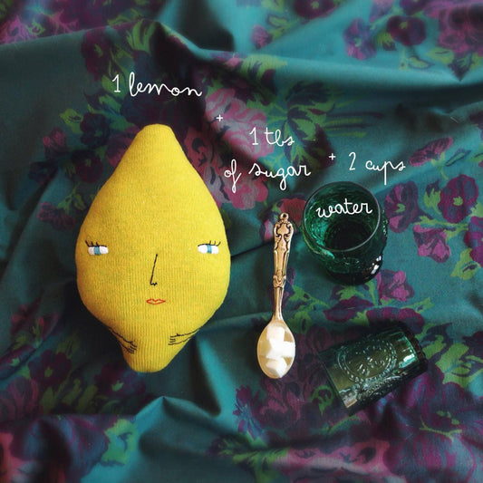 Knitted lemon with face, sugar, water, and spoon on floral tablecloth fabric