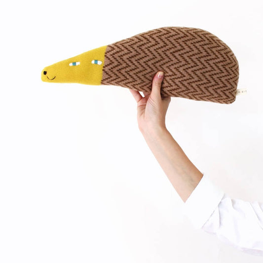 Hand holding a knitted lambswool plush toy shaped like a hedgehog with a brown back and yellow face on a white background