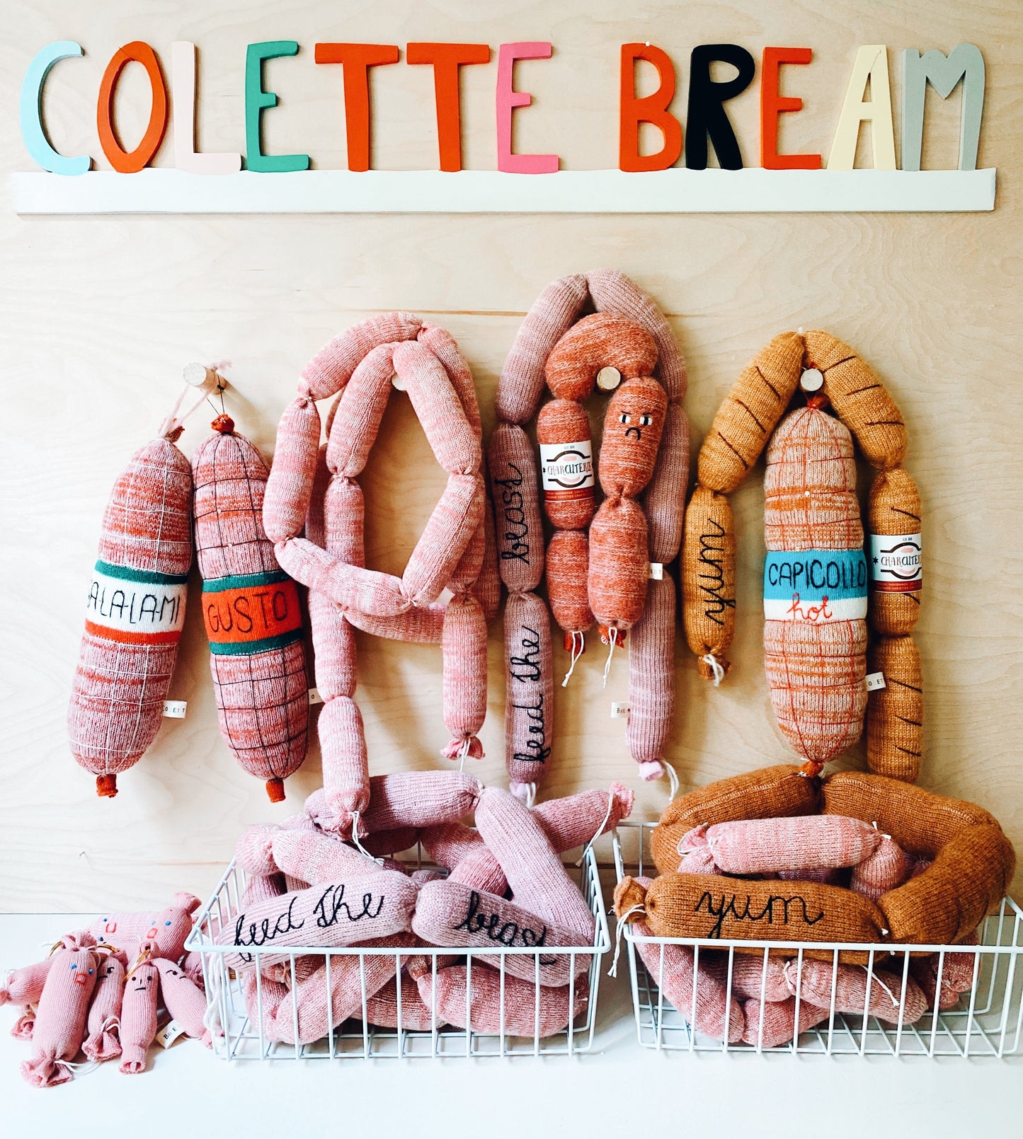 Colorful knitted plush toy sausages and salami with text labels on a white wall with 'COLETTE BREAM' sign.