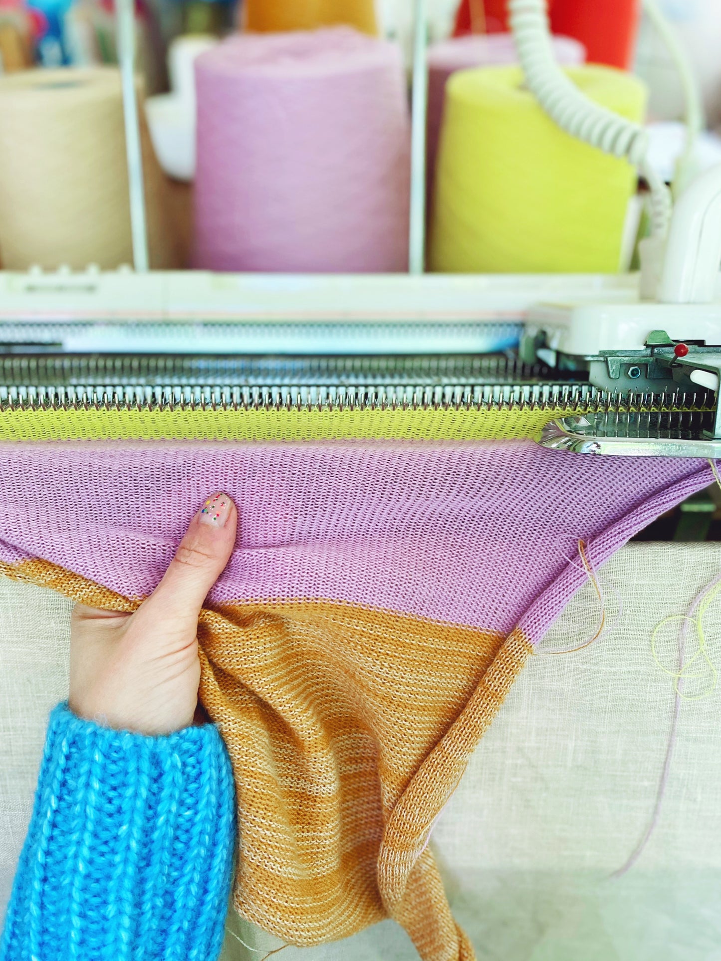 Knitted fabric with a gradient from purple to orange held by a person wearing a blue sweater, with knitting equipment in the background.
