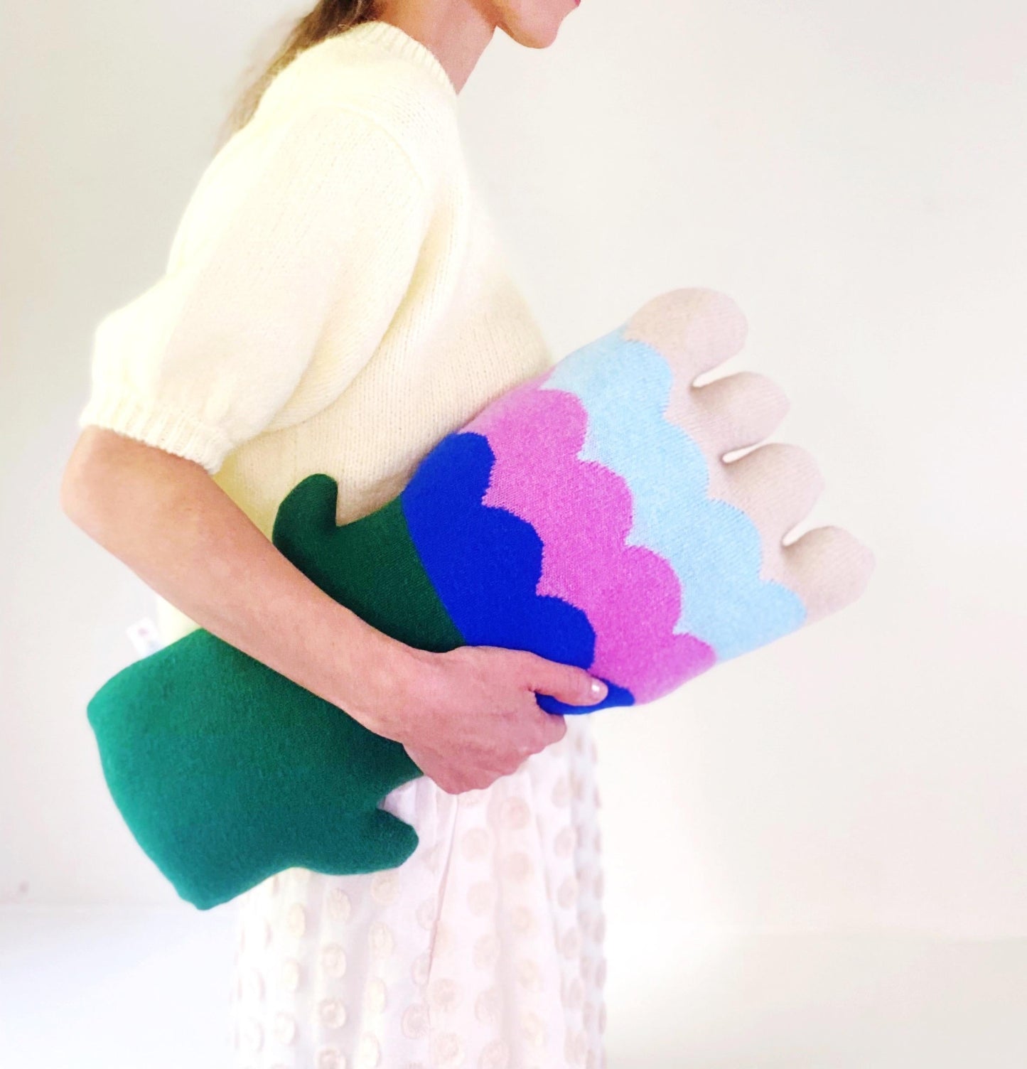 Person holding a colorful knitted flower shaped pillow with blue, pink, and green sections against a white background