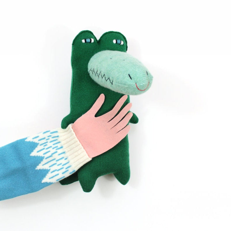 Green knitted plush toy crocodile alligator with a hand holding it on a white background