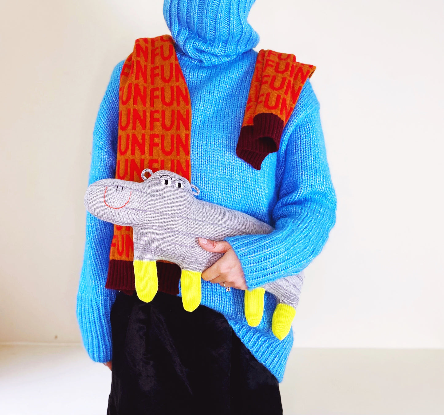 Person wearing a blue sweater with a colorful scarf and holding a knitted hippo plush toy against a white background