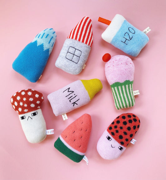 Colorful knitted baby rattle plush toys shaped like bottles, cups, and fruits on a pink background