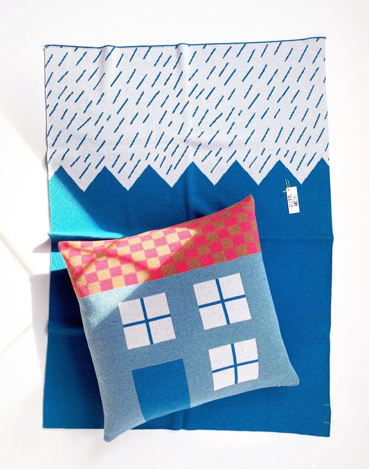Blue knit blanket with geometric mountain design and a colorful house pillow on a white background