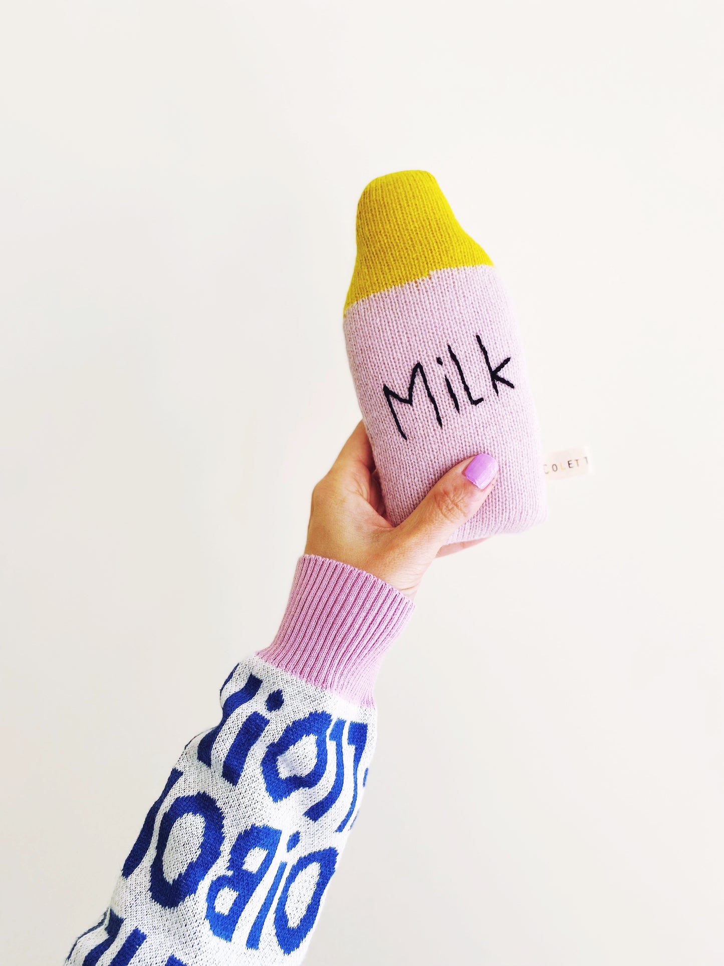 Hand holding a pink knit baby rattle toy shaped like a bottle labeled 'Milk' against a white background