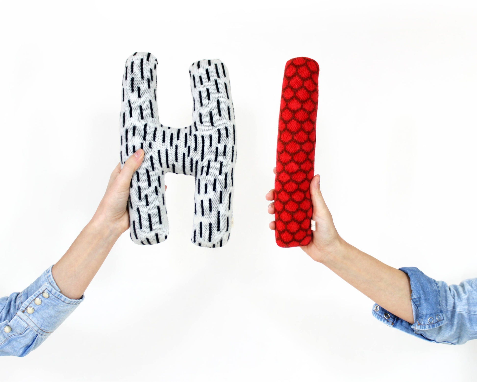 Two hands holding letter 'H' and 'I' shaped pillows that together spell 'Hi", one black and white patterned, one red.