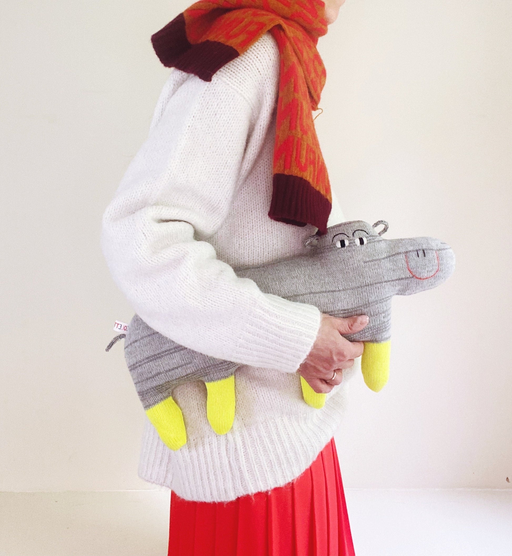 Person wearing a white sweater with a red scarf, holding a gray knitted hippo toy with yellow accents.