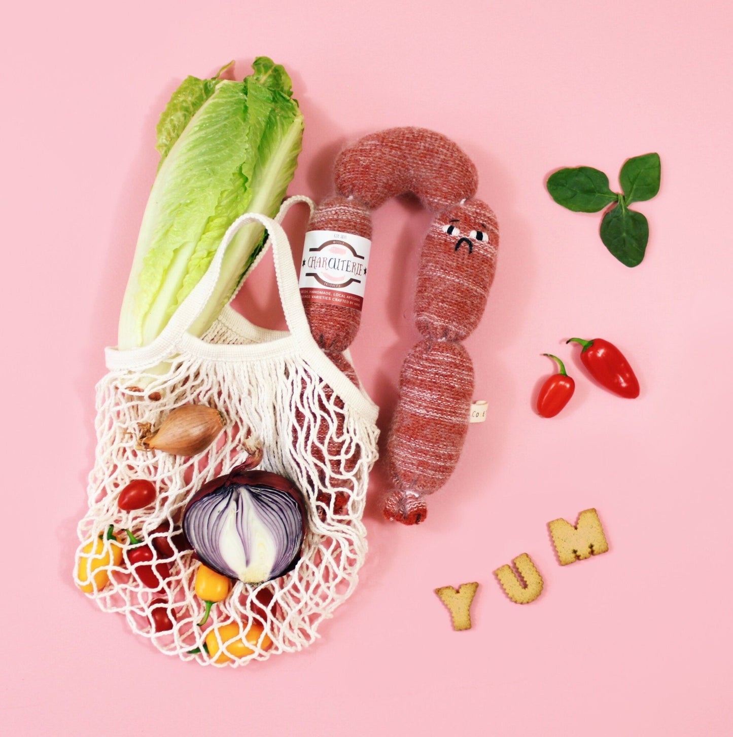 Mesh bag with play food groceries, salami, knitted sausage and vegetables on a pink background with 'YUM' letters.