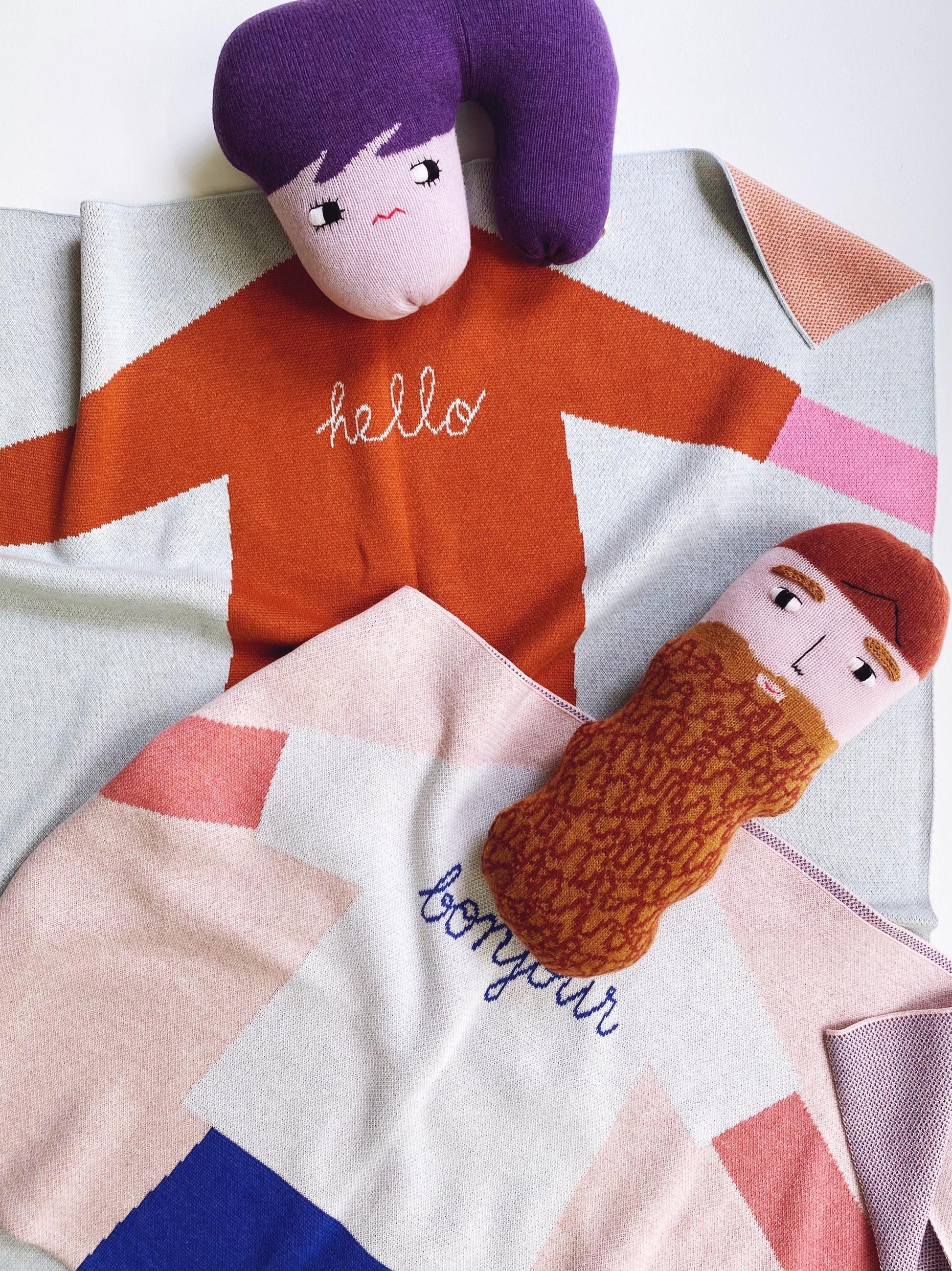 Two plush toys on a geometric-patterned blanket with 'hello' and 'bonjour' text.
