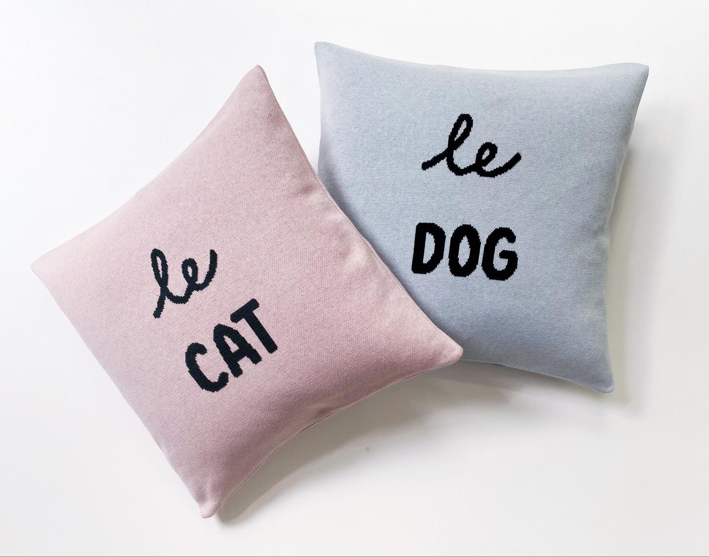 Two knitted cushions, one pink and one gray, with 'le cat' and 'le dog' text on a white background.