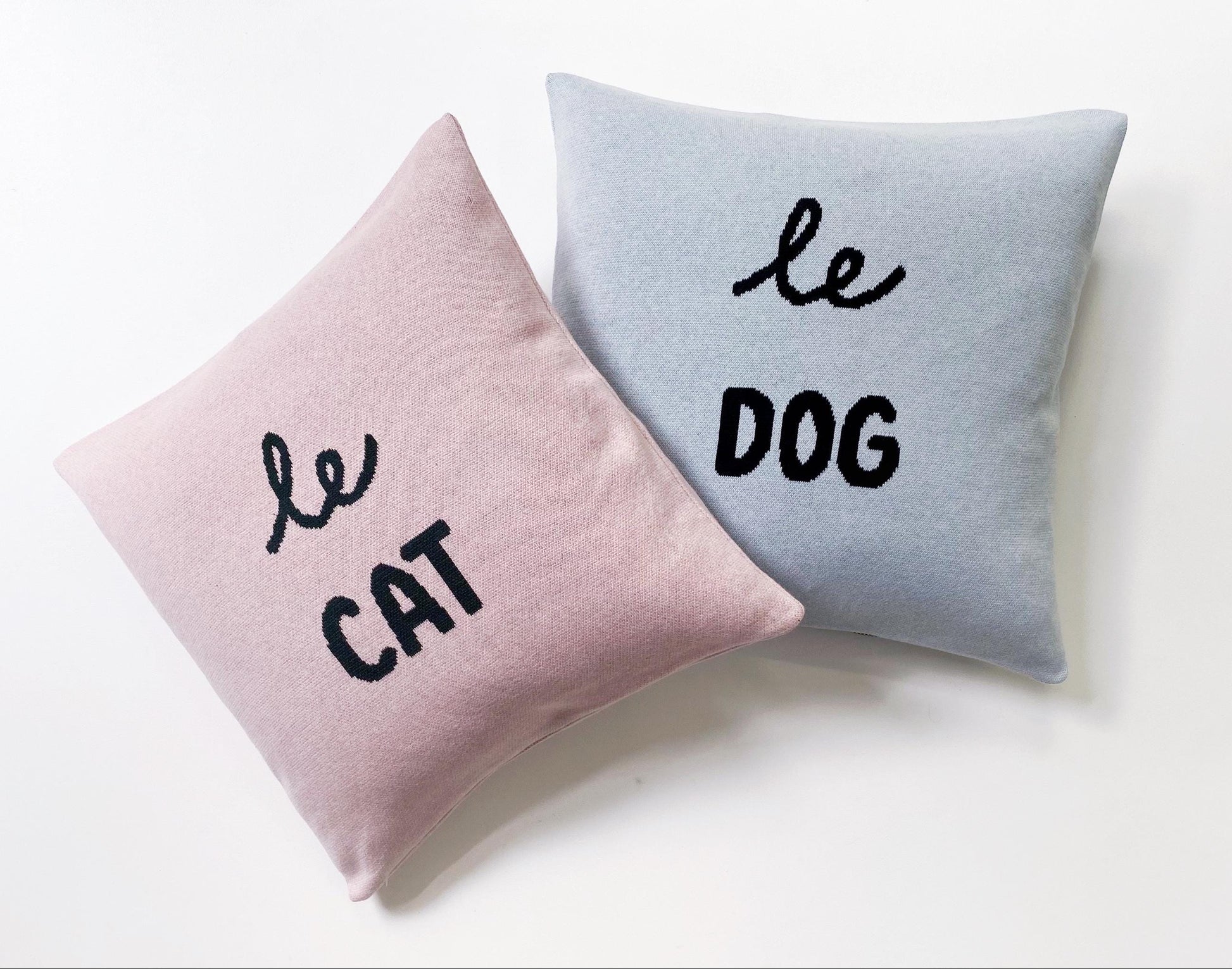 Two knitted cushions, one pink and one gray, with 'le cat' and 'le dog' text on a white background.