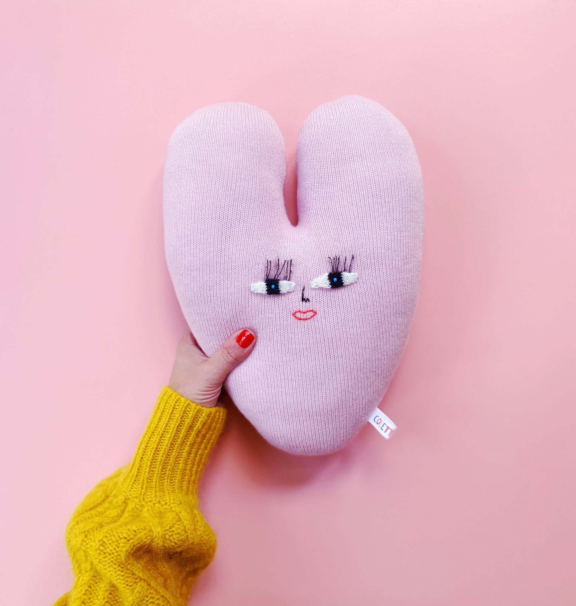 Pink heart-shaped knitted pillow with a face design held by a hand against a pink background