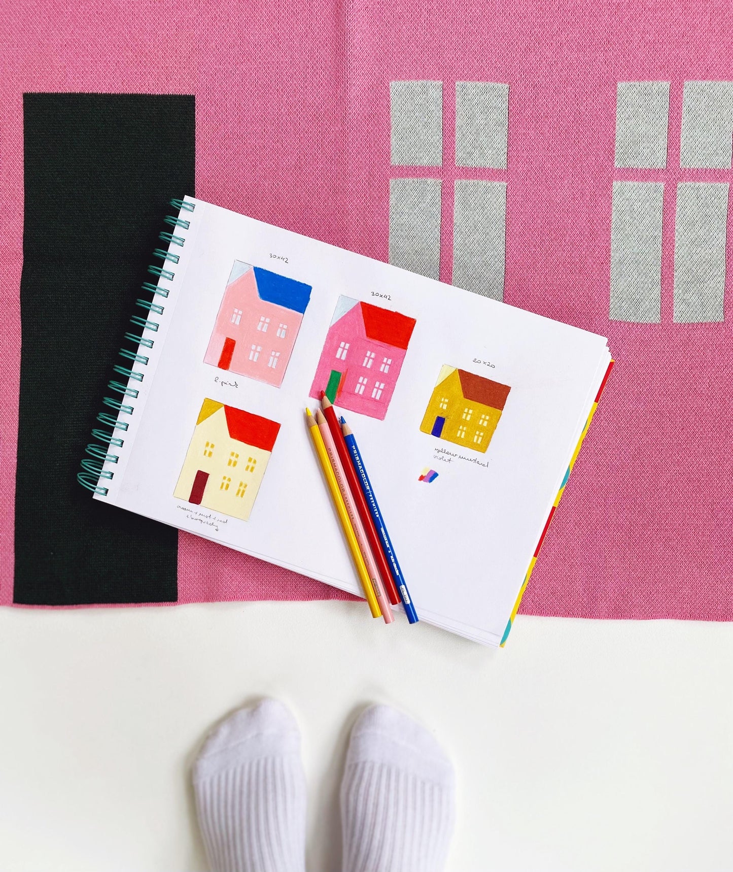 Notebook with house illustrations, colored pencils, and socks on a pink knitted blanket 