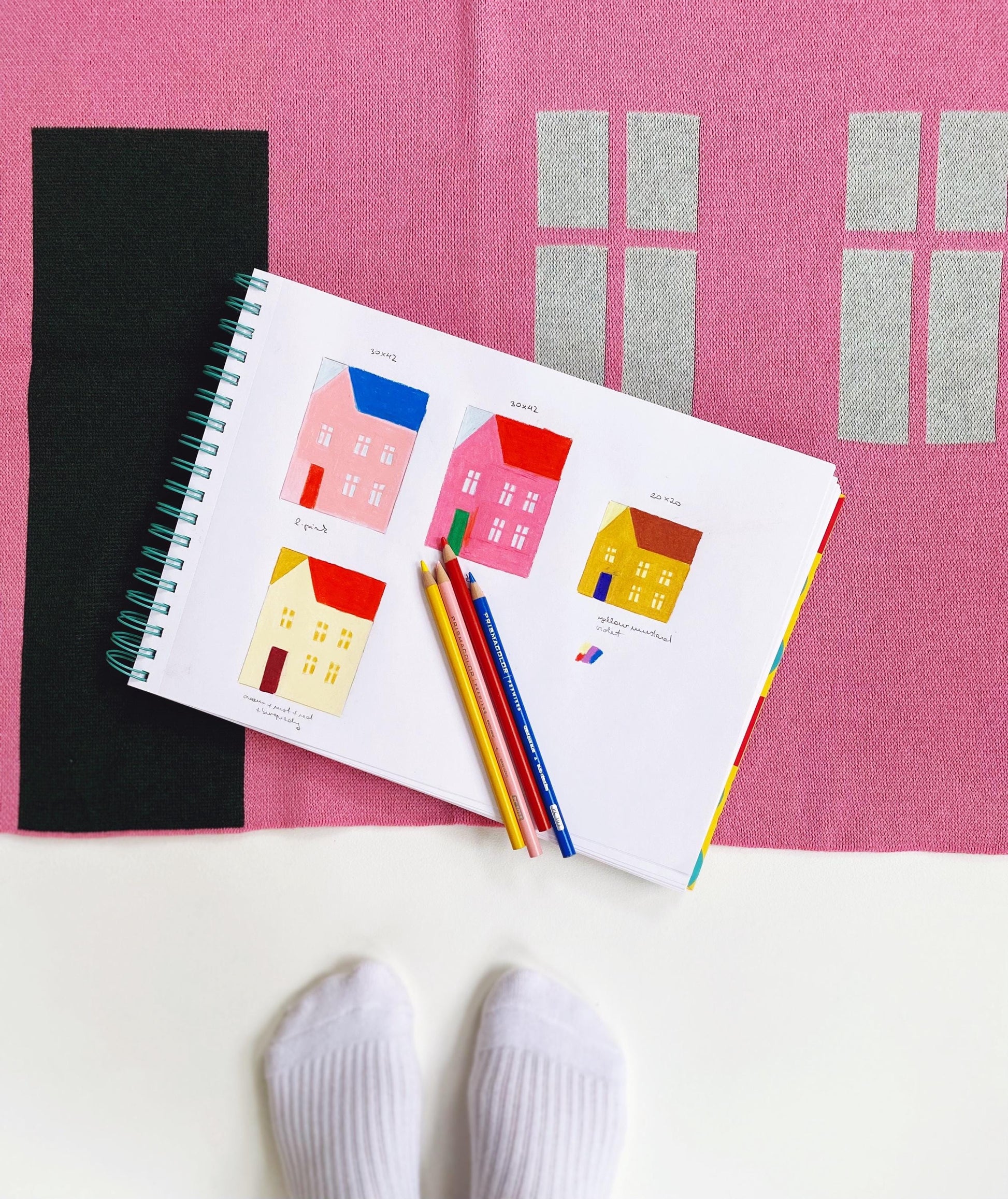 Notebook with house illustrations, colored pencils, and socks on a pink knitted blanket 