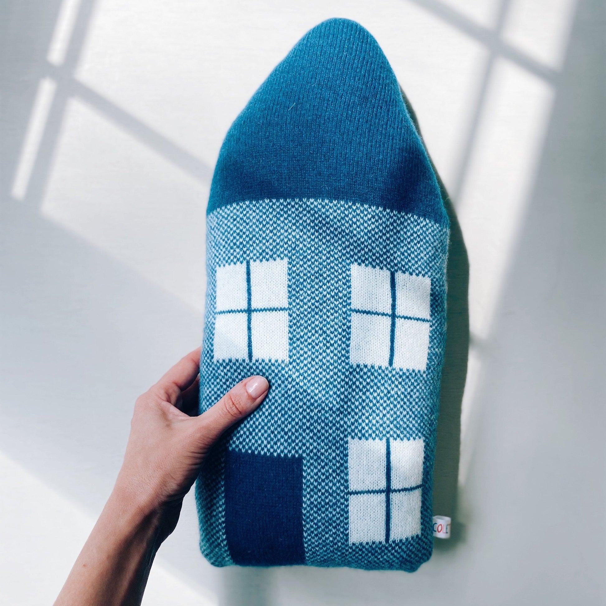Blue knitted house-shaped pillow with white windows held by a hand against a light background