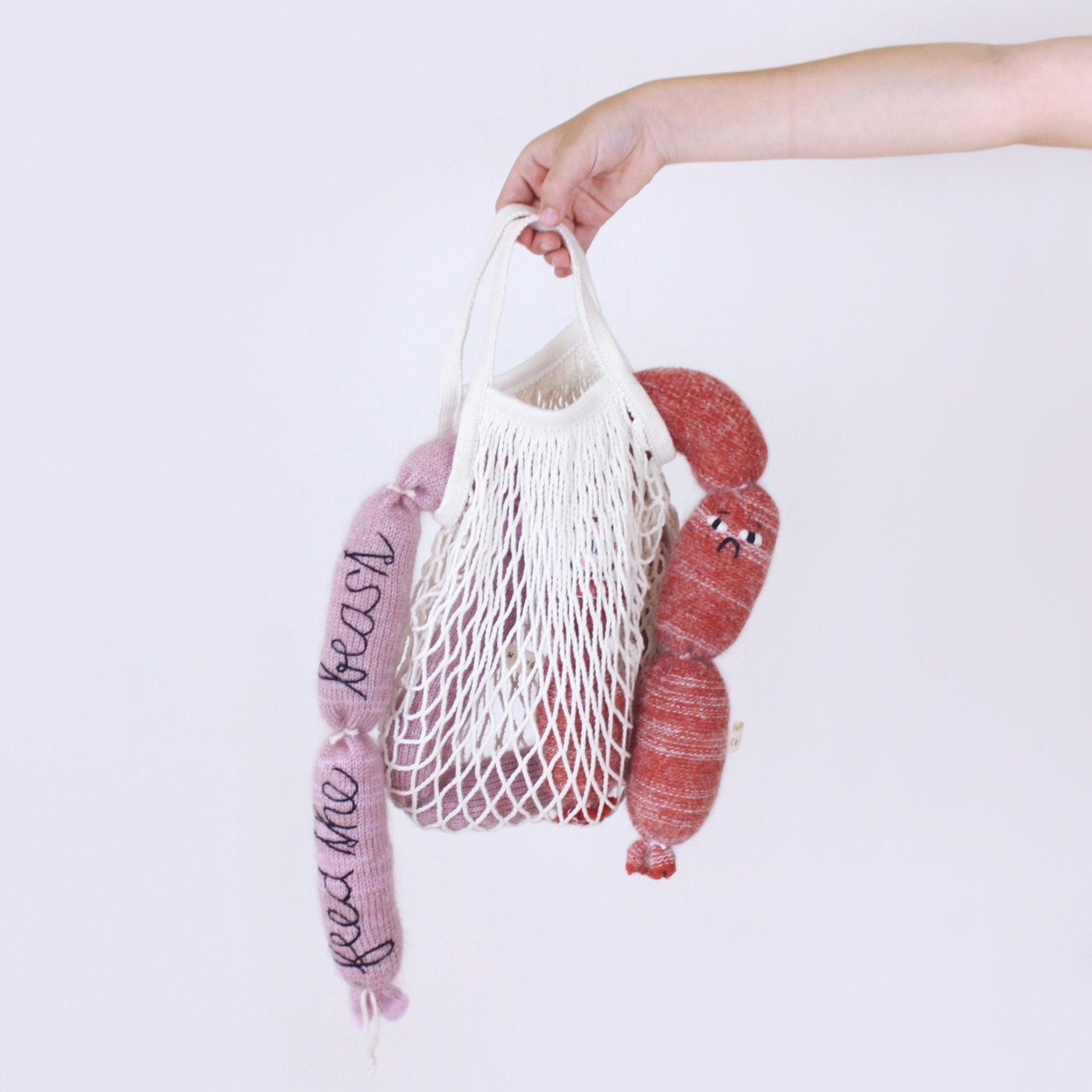 Hand holding a white mesh bag with pink and red knitted sausages against a light purple background