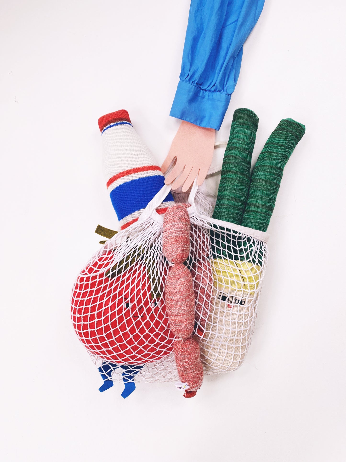 Person holding a mesh bag with groceries including a bottle, vegetables, and a sausage on a white background