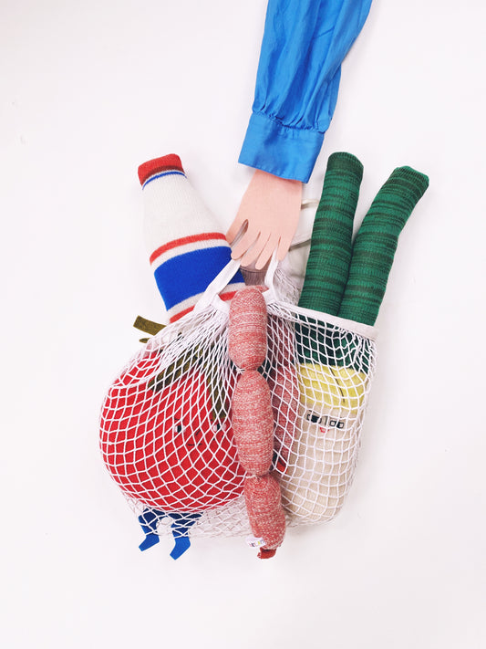 Person holding a mesh bag with groceries including a bottle, vegetables, and a sausage on a white background