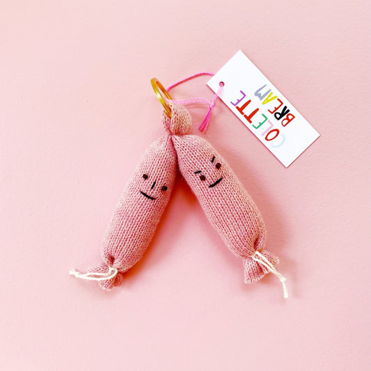 Two pink knitted sausages keychains with faces on a pink background, featuring a colorful tag.