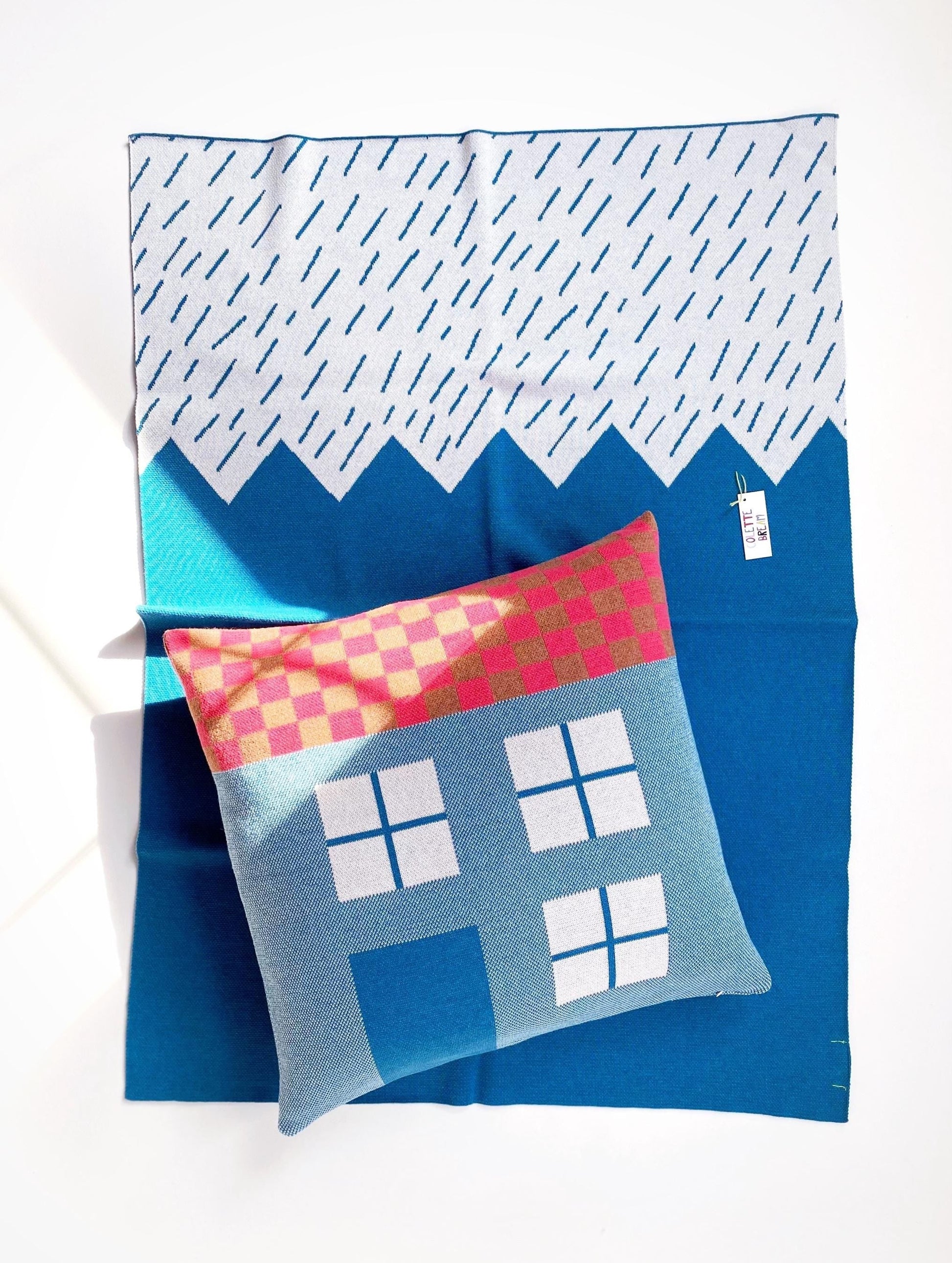 Knit house pillow with colorful checkered pattern and a blue blanket with mountain design on a white background