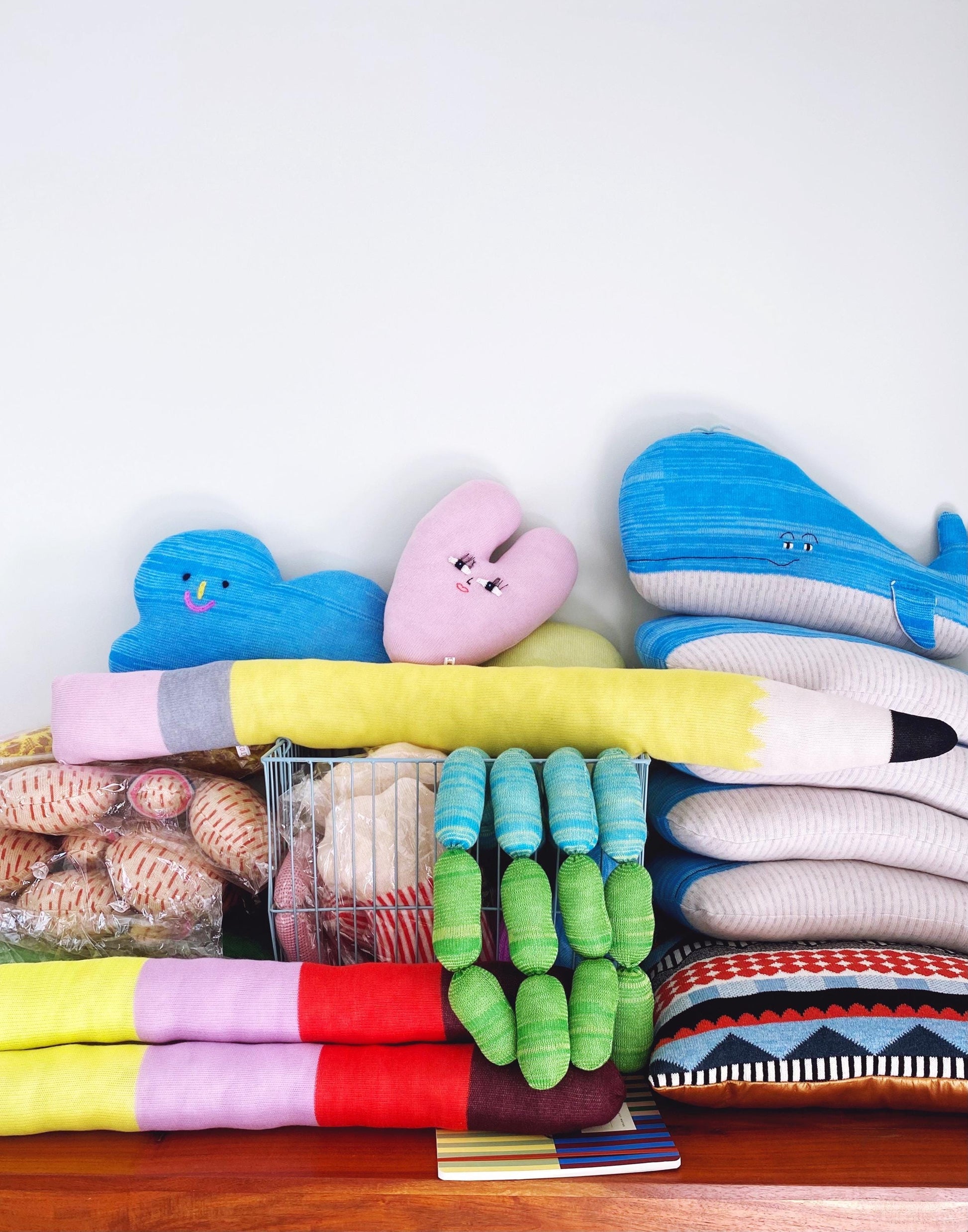 Stack of colorful knitted plush toys and pillows on a wooden surface with a white background