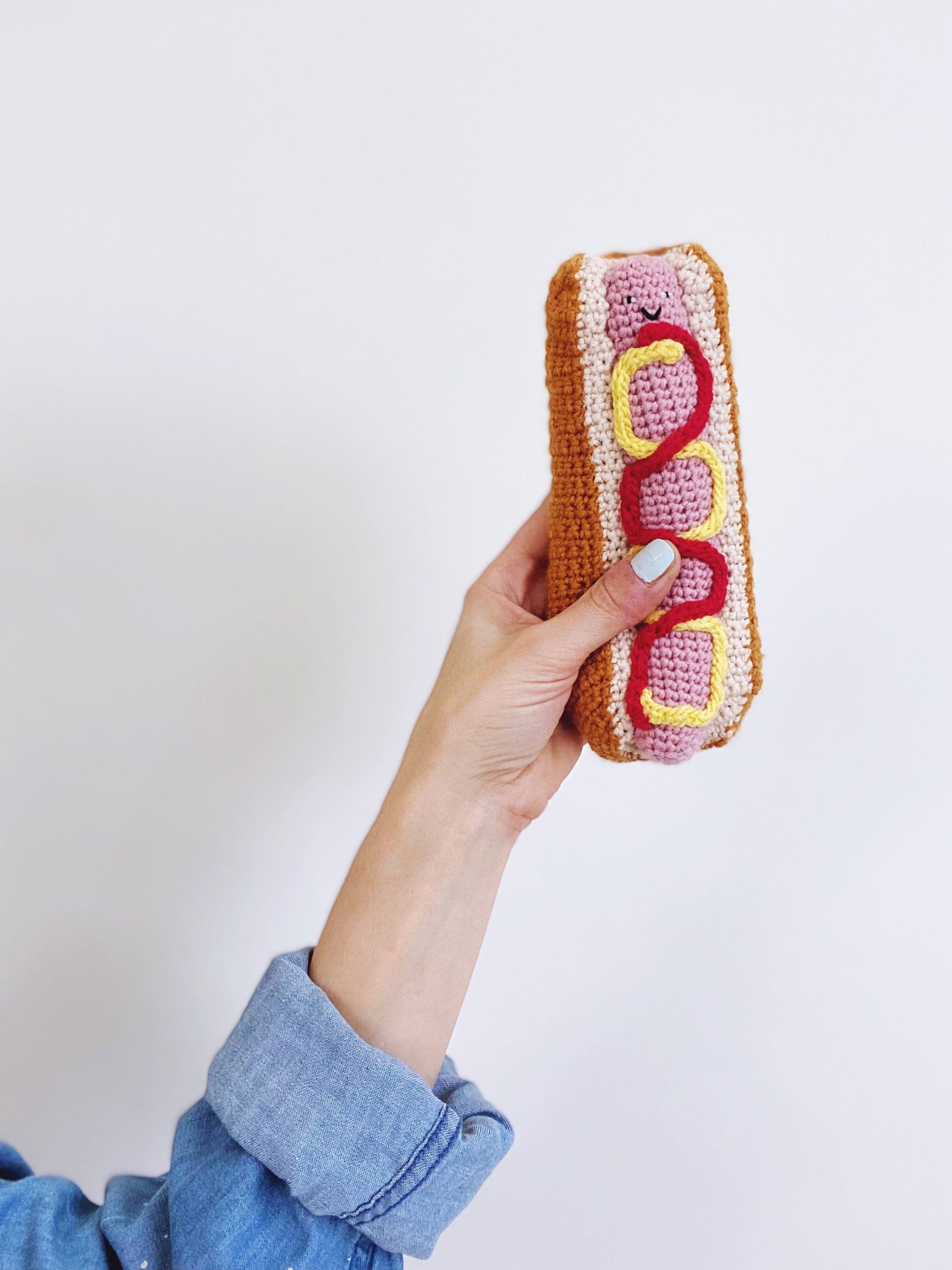 Hand holding a crocheted hot dog toy against a white background