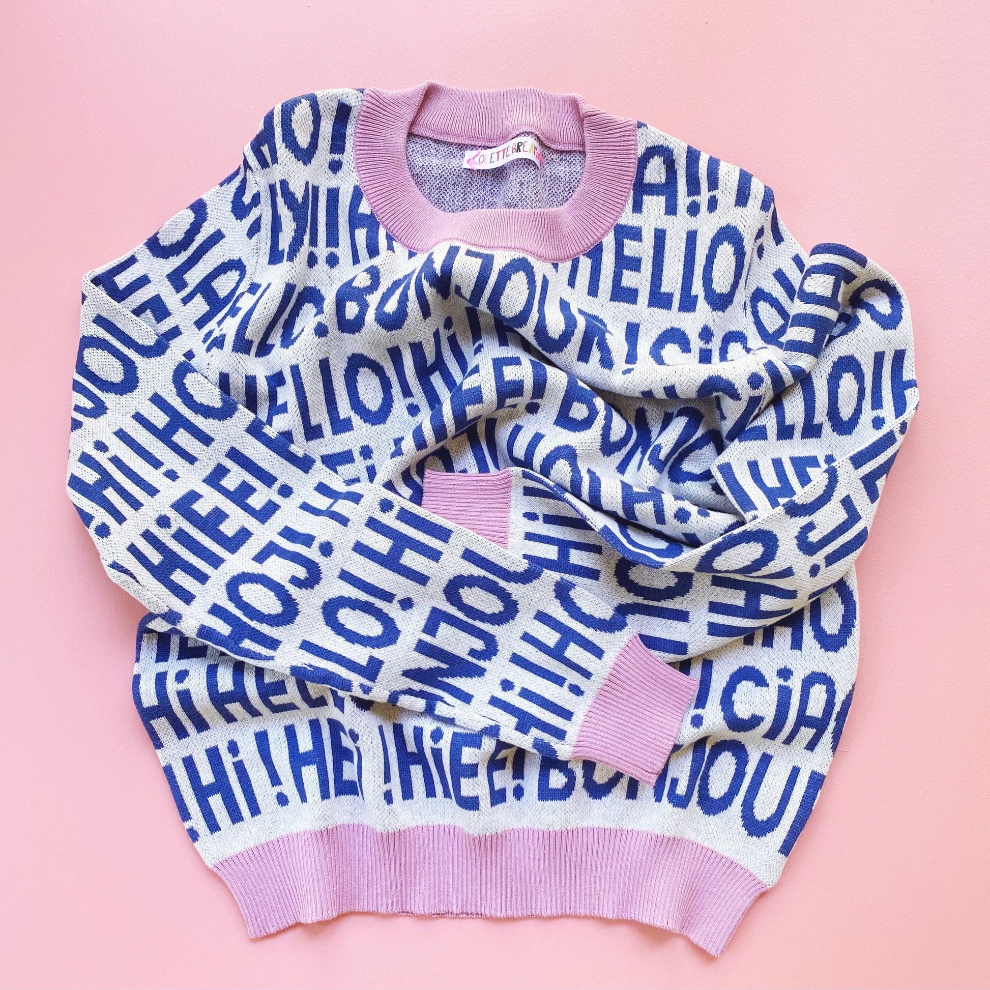 Knit white and blue sweater with 'HELLO!' greetings text on a pink background