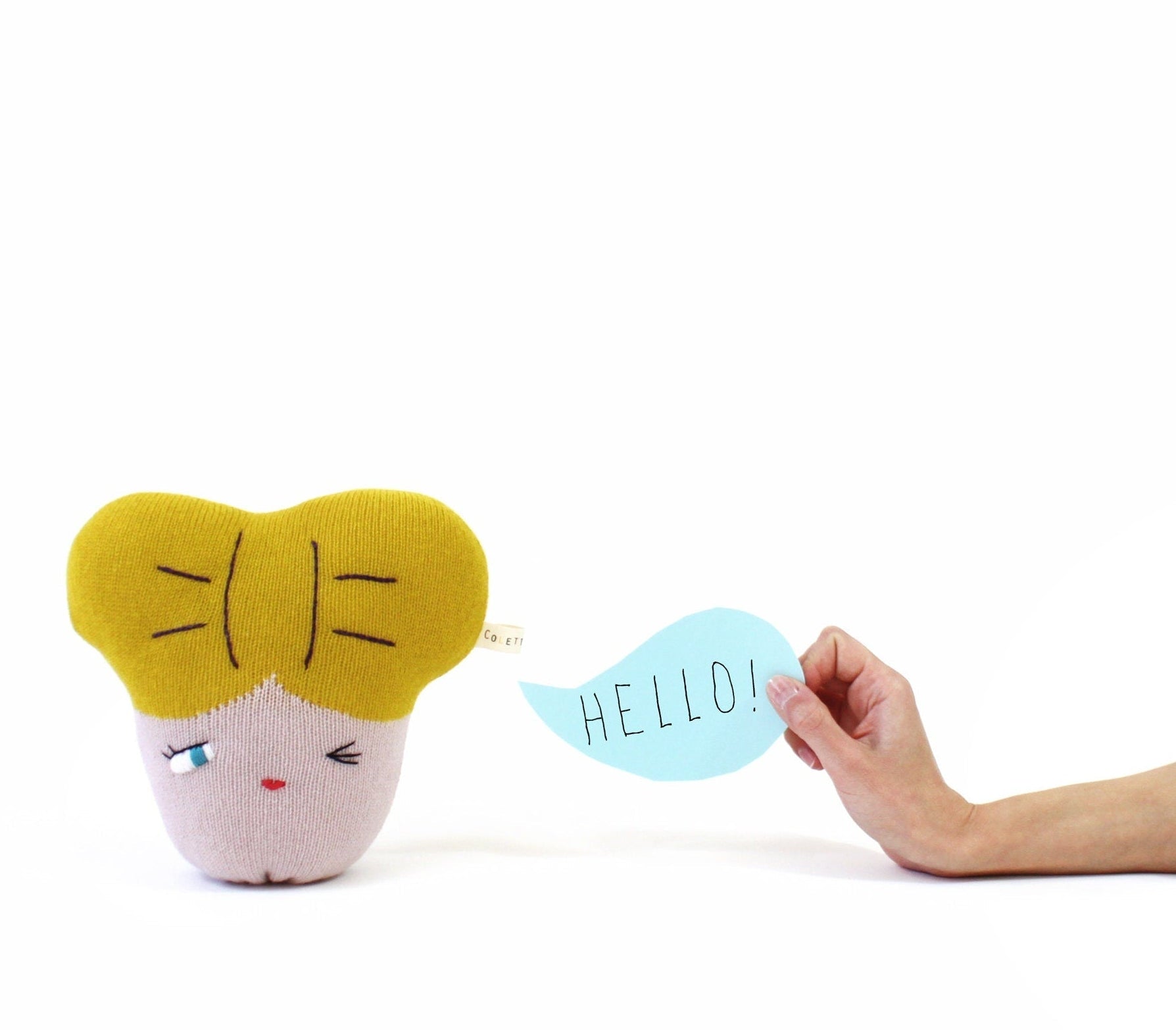 Small knitted plush toy with a yellow hair bow and pink face holding a 'HELLO!' sign against a white background