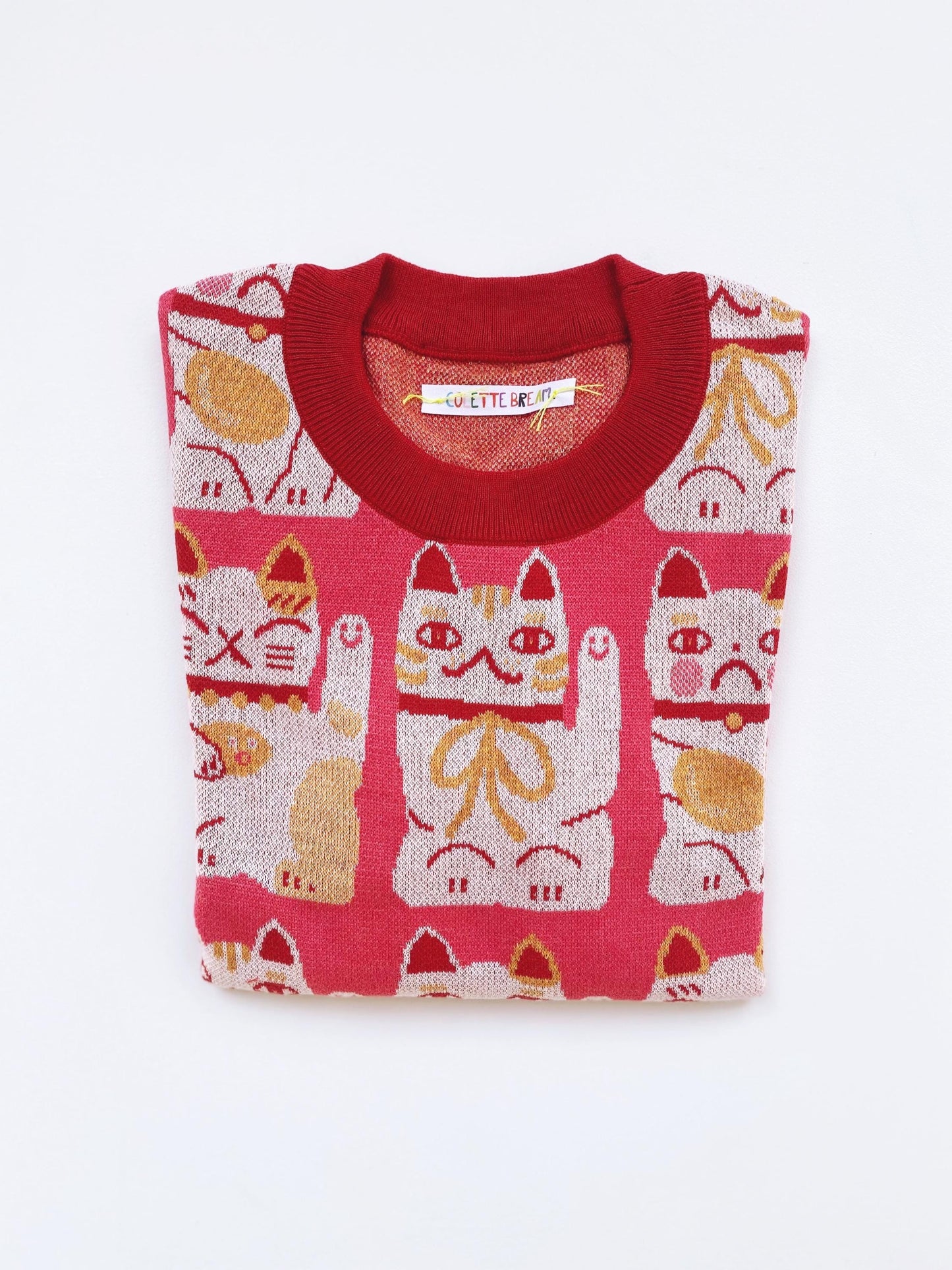 Folded red and pink knitted sweater with maneki neko cat pattern on a white background