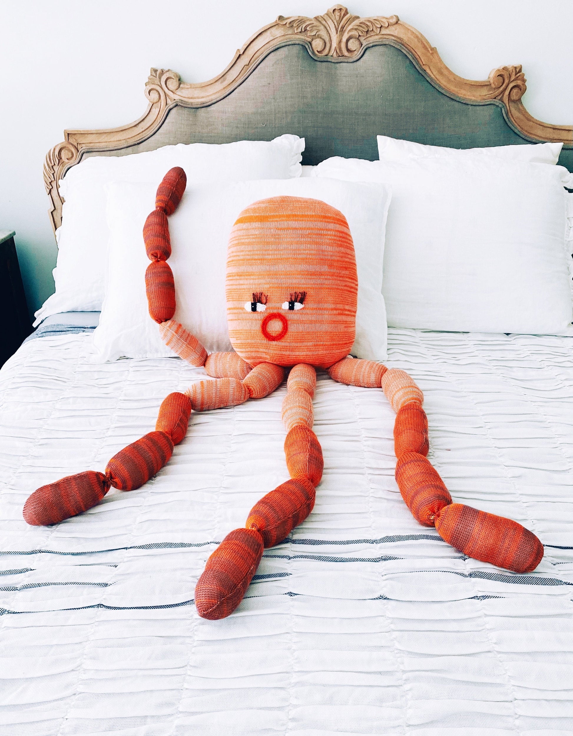 Large knitted plush toy resembling a giant octopus character on a bed with white pillows and a decorative headboard.