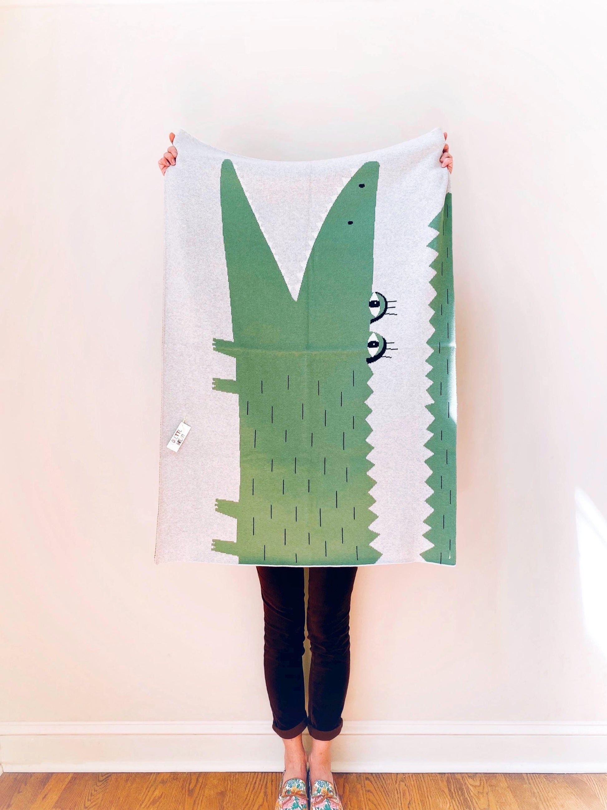 Person holding a knit baby blanket with alligator design against a white wall.