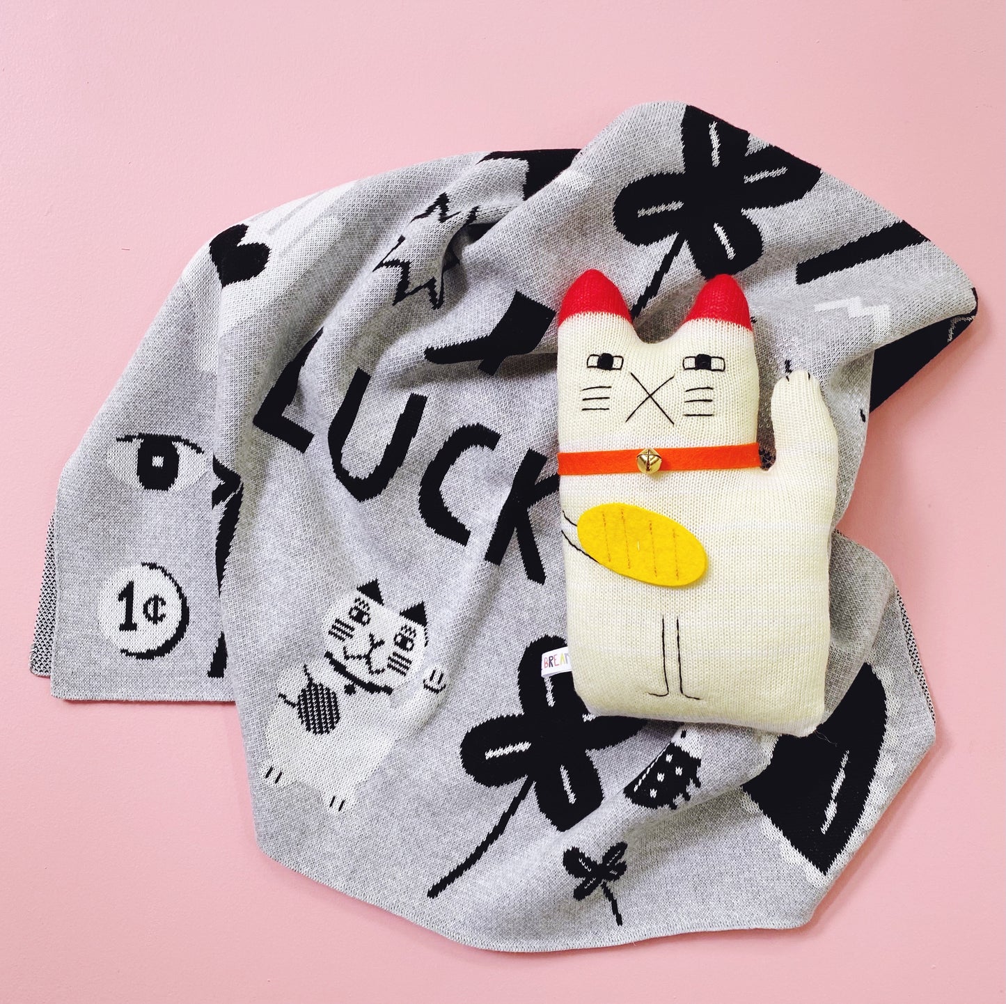 Cat-shaped plush toy on a gray blanket with lucky charms designs on a pink background