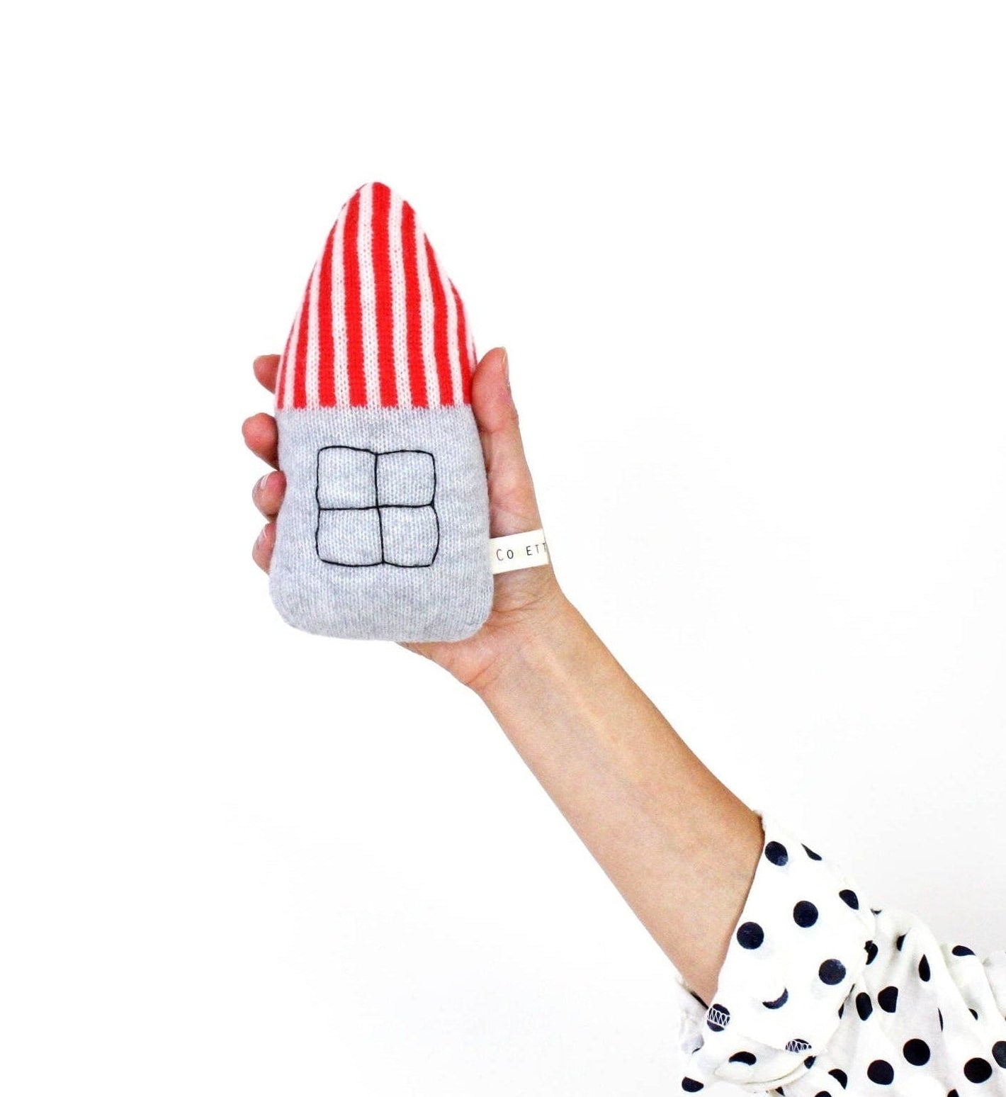 Hand holding a small house-shaped baby rattle toy with red and white striped roof and gray body on a white background