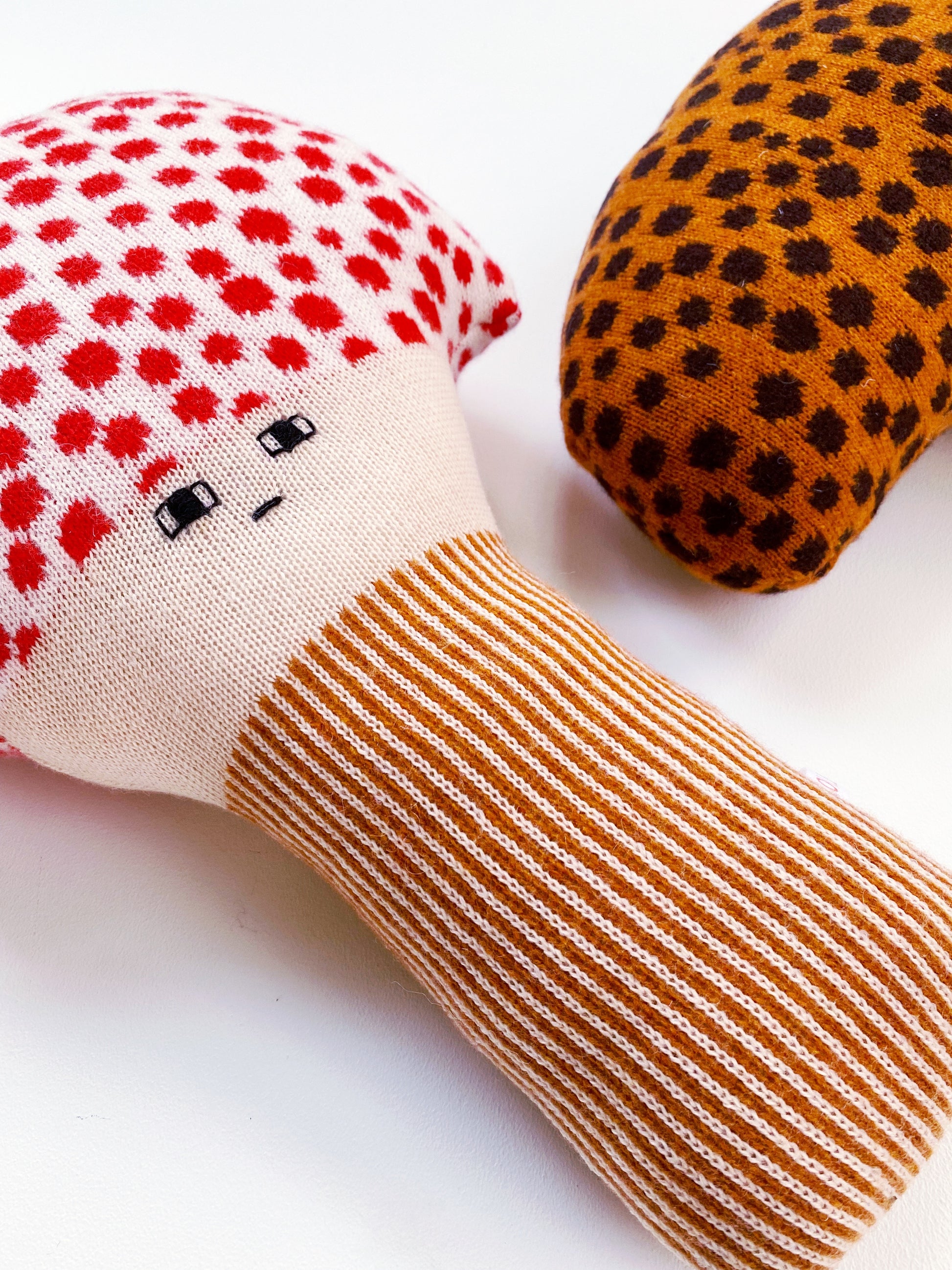 Knitted mushroom toy with a face, red and white polka dot top, brown handle, and orange and black polka dot top on a white background.