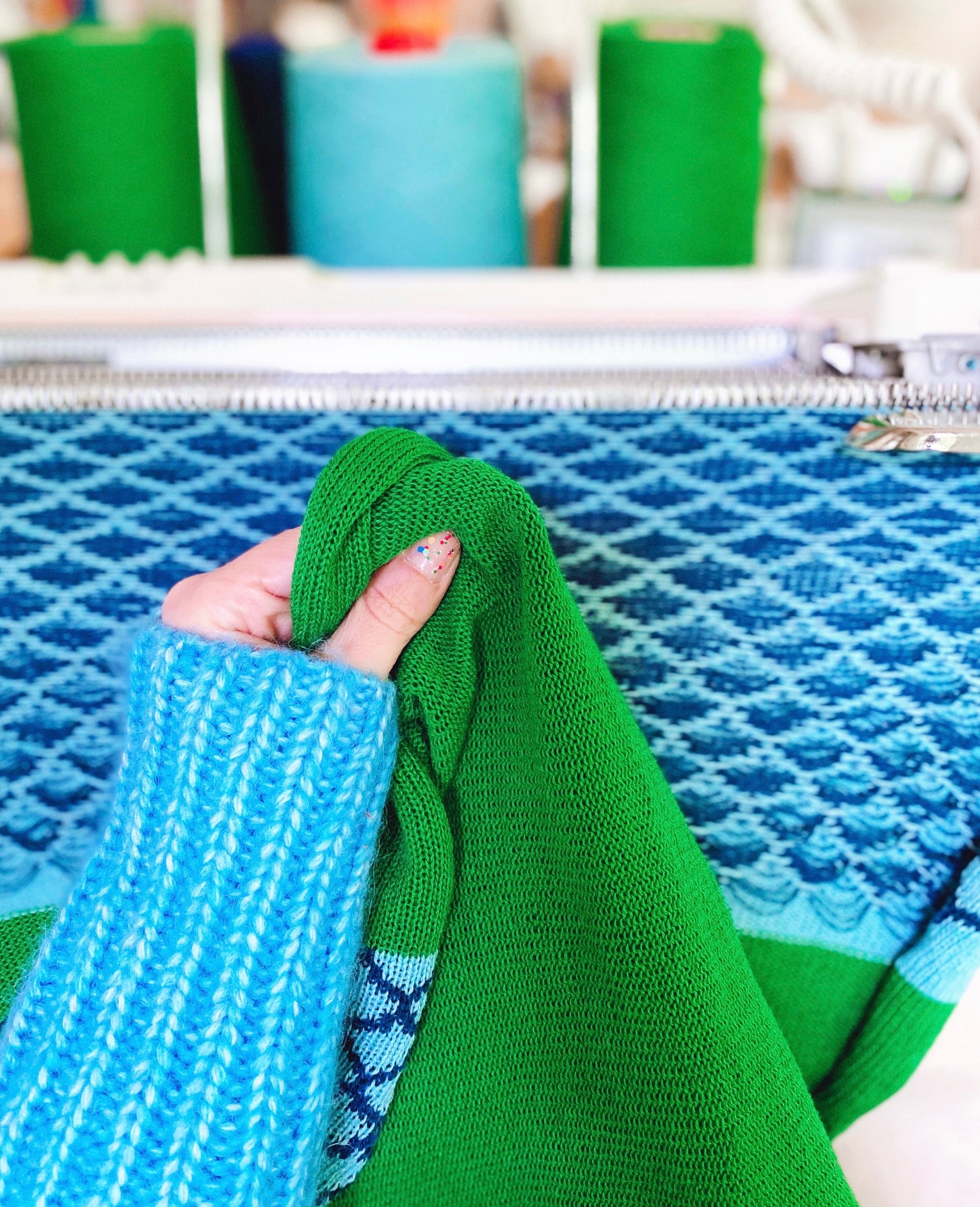 Hand holding green fabric against a blue textured surface with spools of thread in the background.