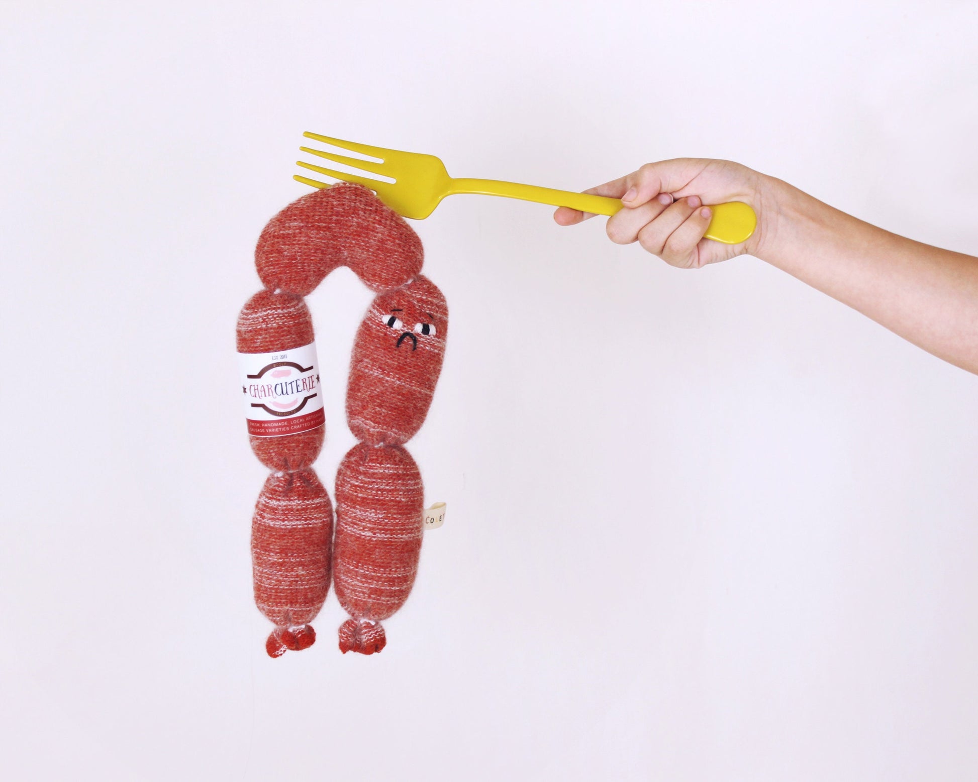 Hand holding a yellow fork over knitted sausages with a face drawn on them against a white background