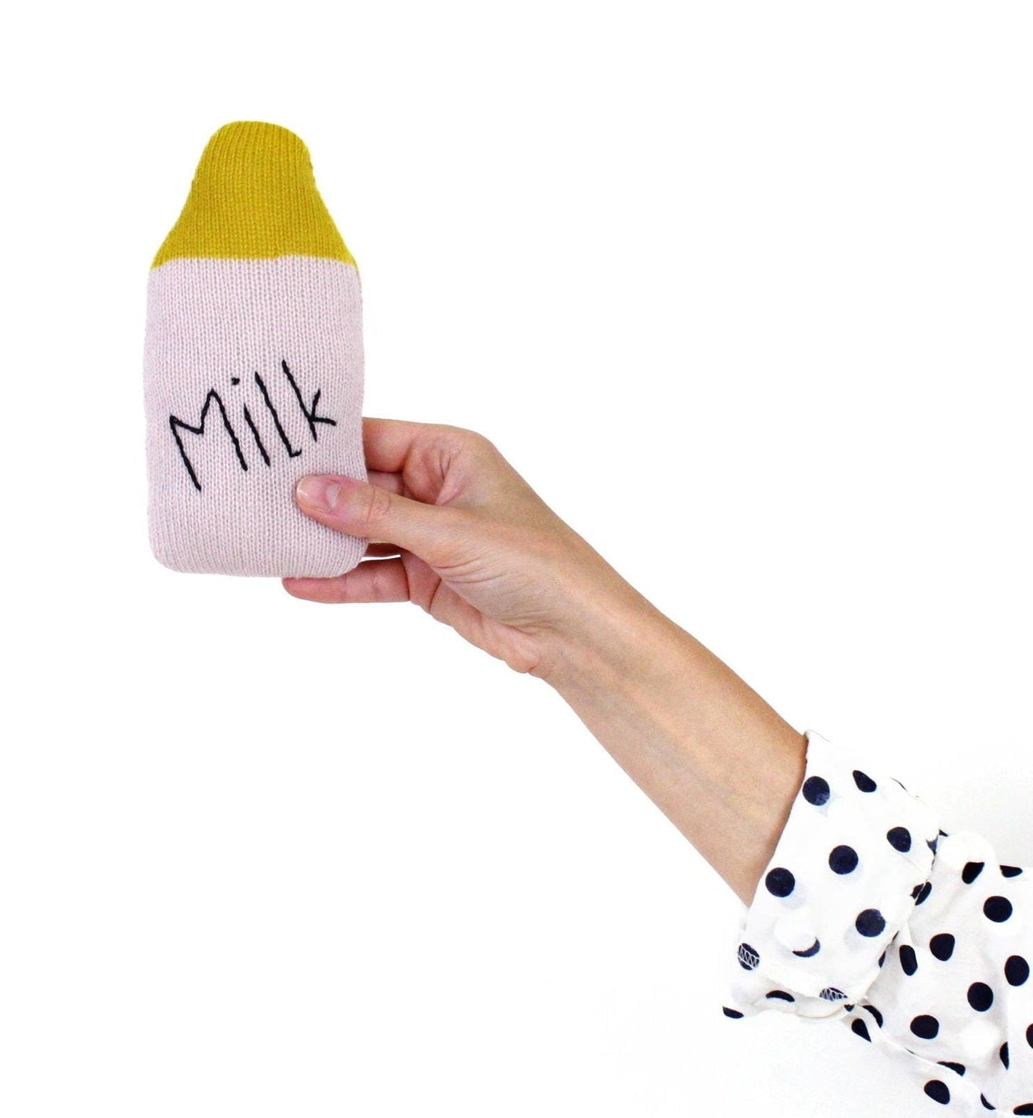 Hand holding a pink and yellow milk bottle-shaped baby rattle plush toy with 'Milk' text on a white background