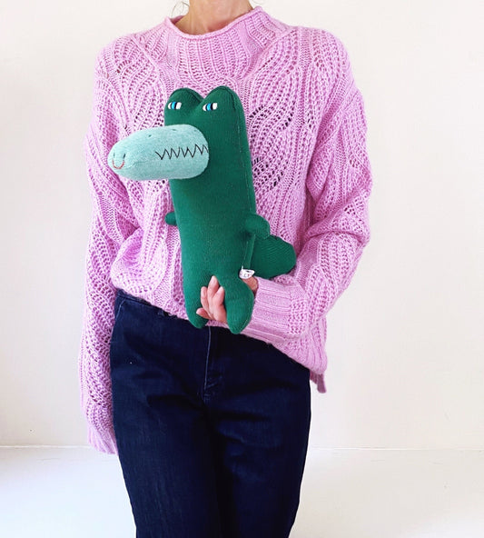 Person wearing a pink sweater holding a green knitted crocodile plush toy 
