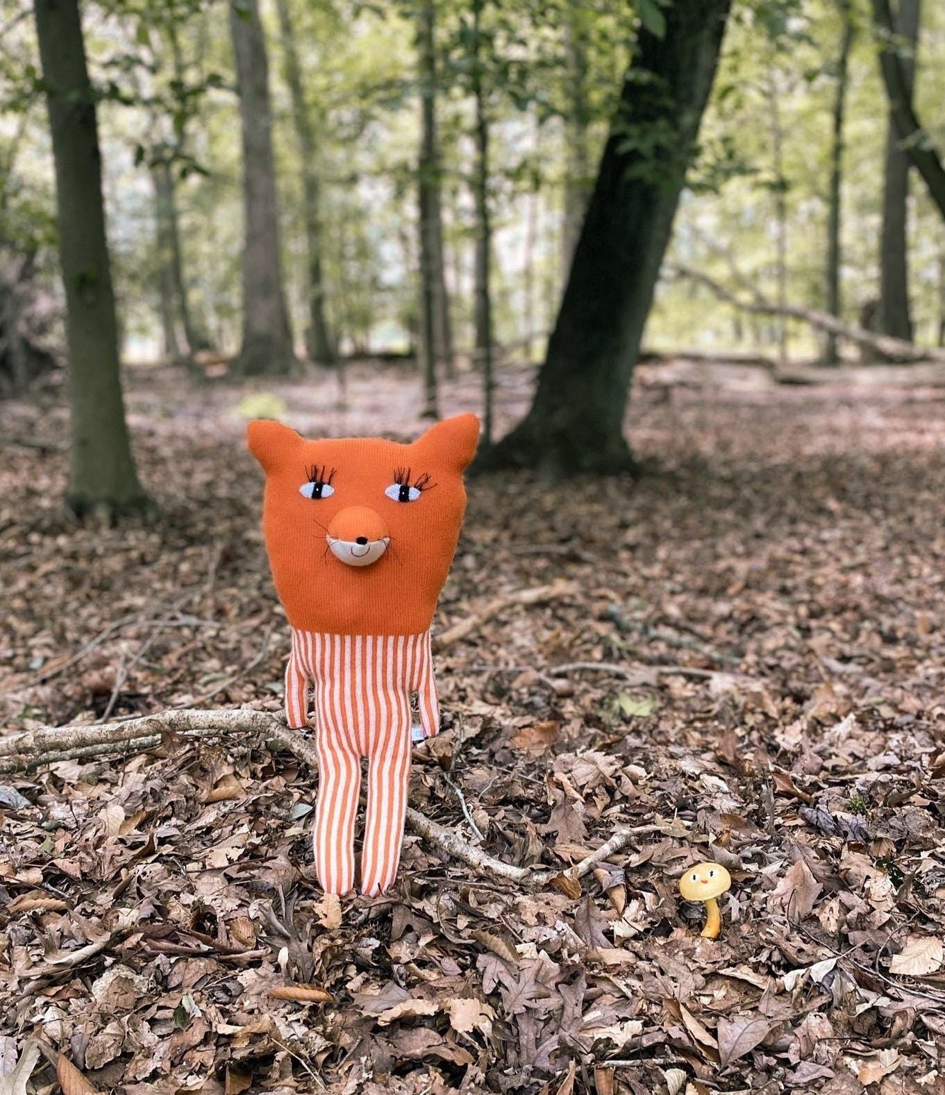 Plush toy fox in a forest setting with trees and fallen leaves.