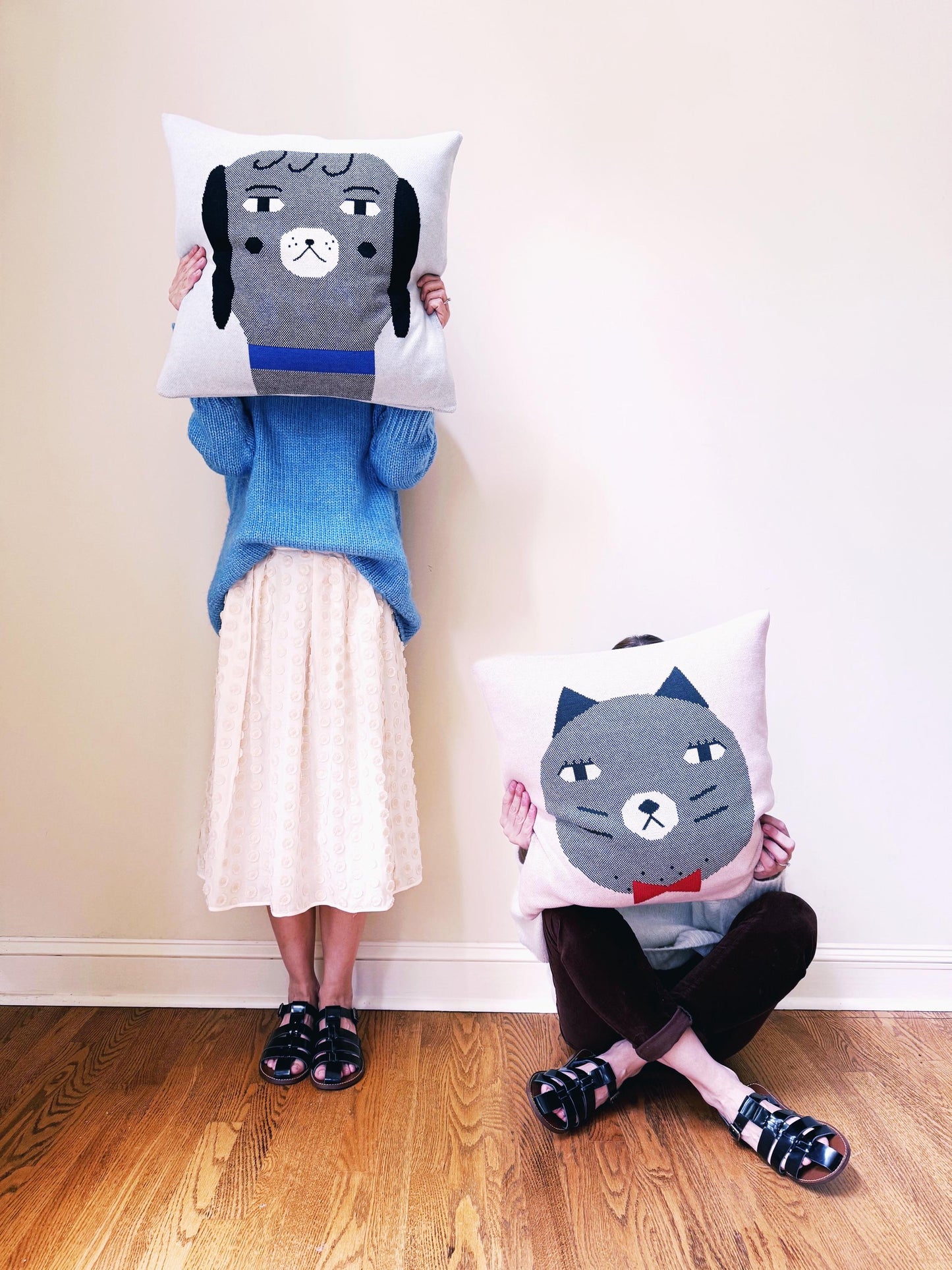 Two people holding cat and dog themed cushions against a plain wall.
