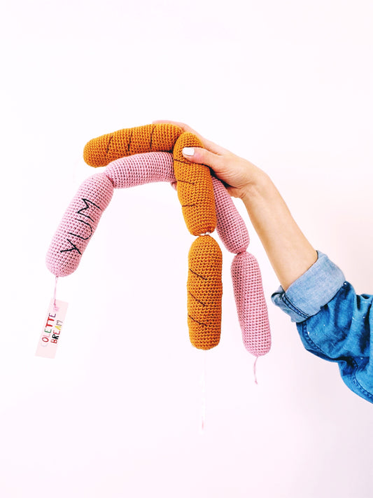 Hand holding a colorful knitted sausage shaped toy with pink, orange, and yellow sections on a white background.