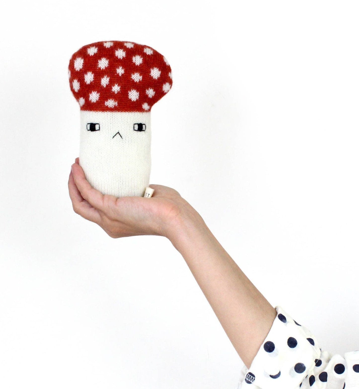 Hand holding a white mushroom baby rattle plush toy with a red polka dot mushroom cap on a white background