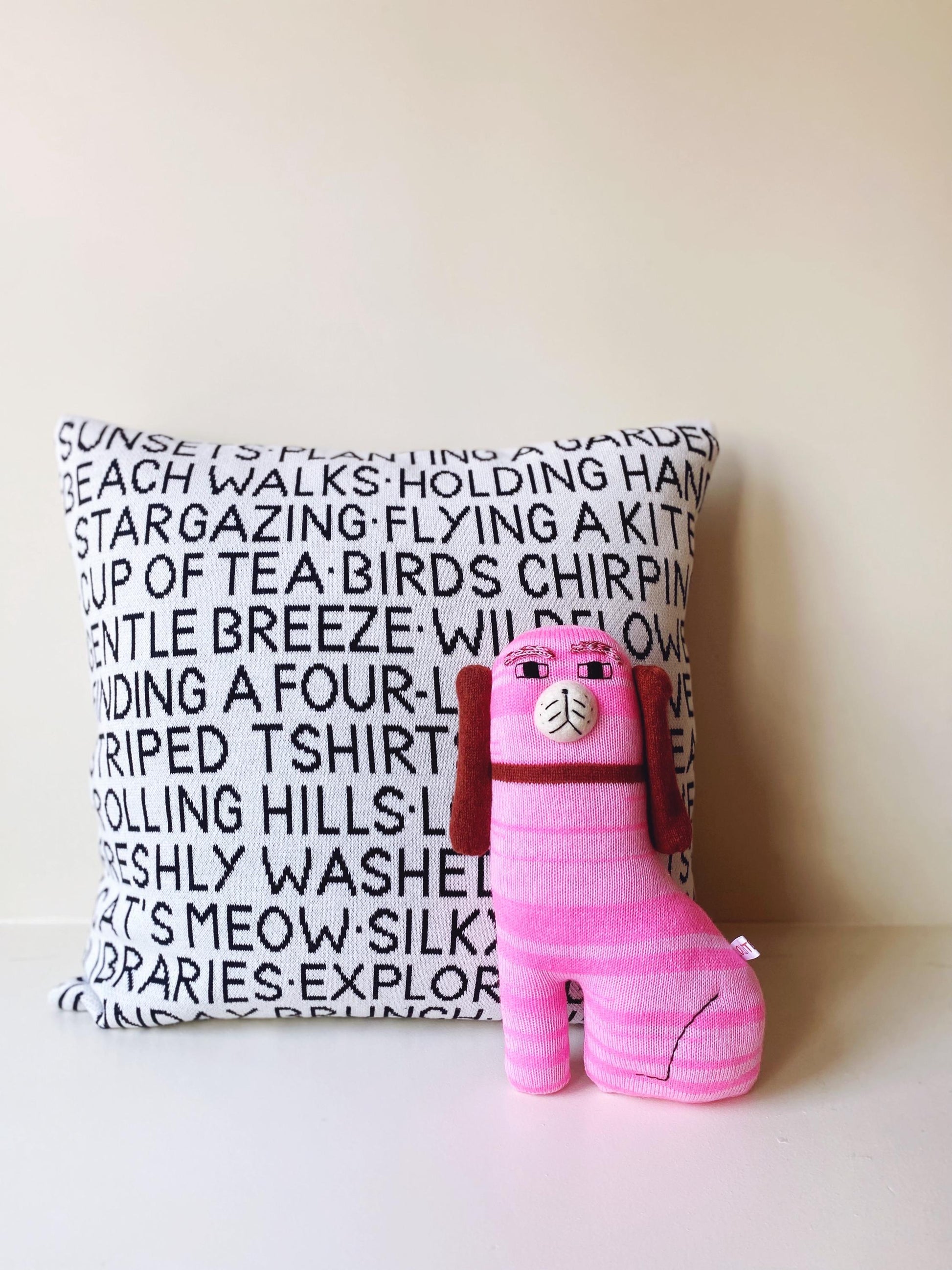 Pink staffordshire dog plush toy sitting on a surface next to a decorative wordy pillow with text.