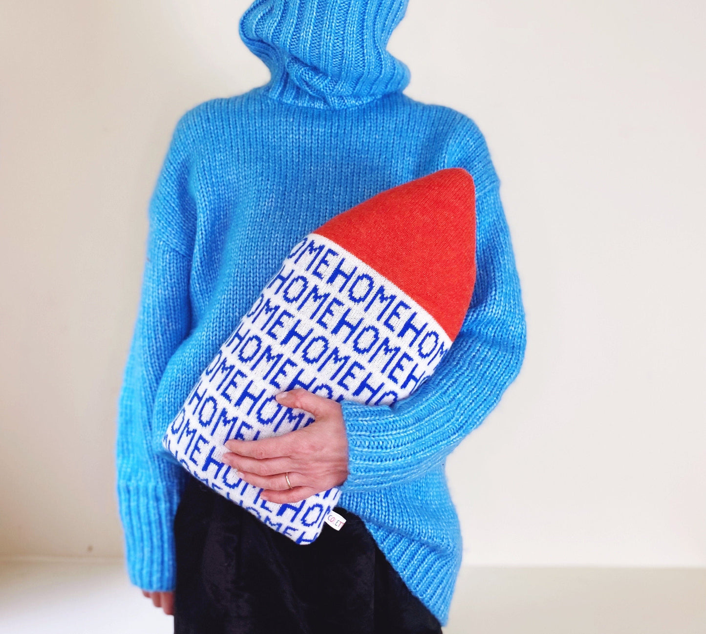 Person wearing a blue sweater holding a red and white house-shaped pillow with 'HOME' text.