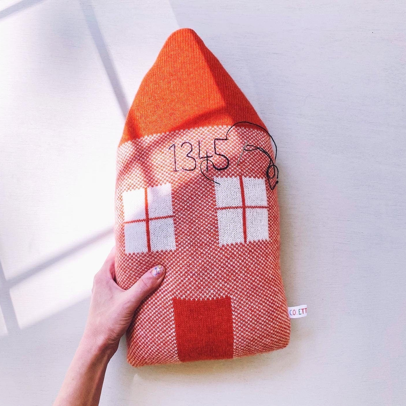 Red and orange house-shaped pillow with a hand holding it on a white background