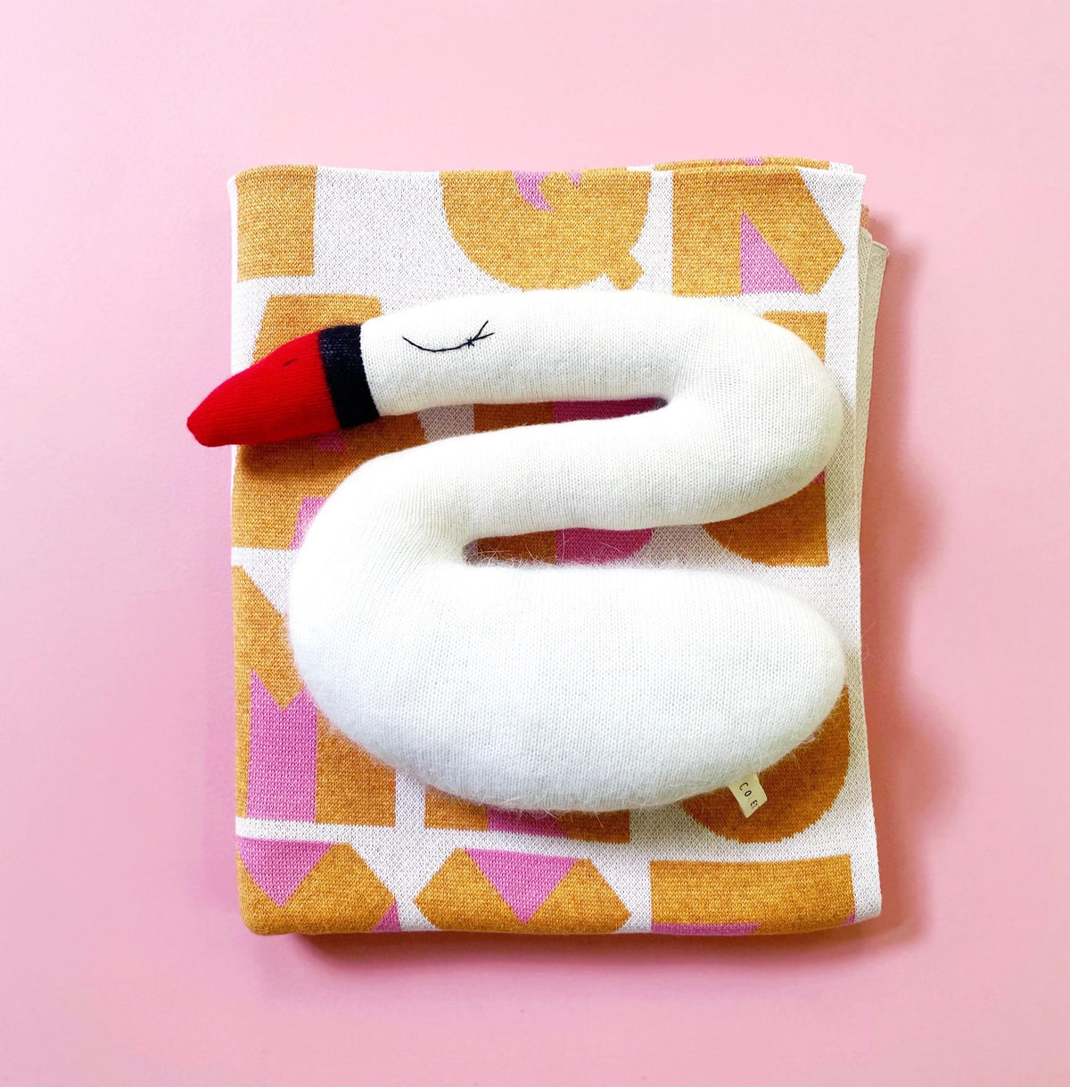 White swan-shaped pillow plush on a folded alphabet patterned knitted blanket with a pink background