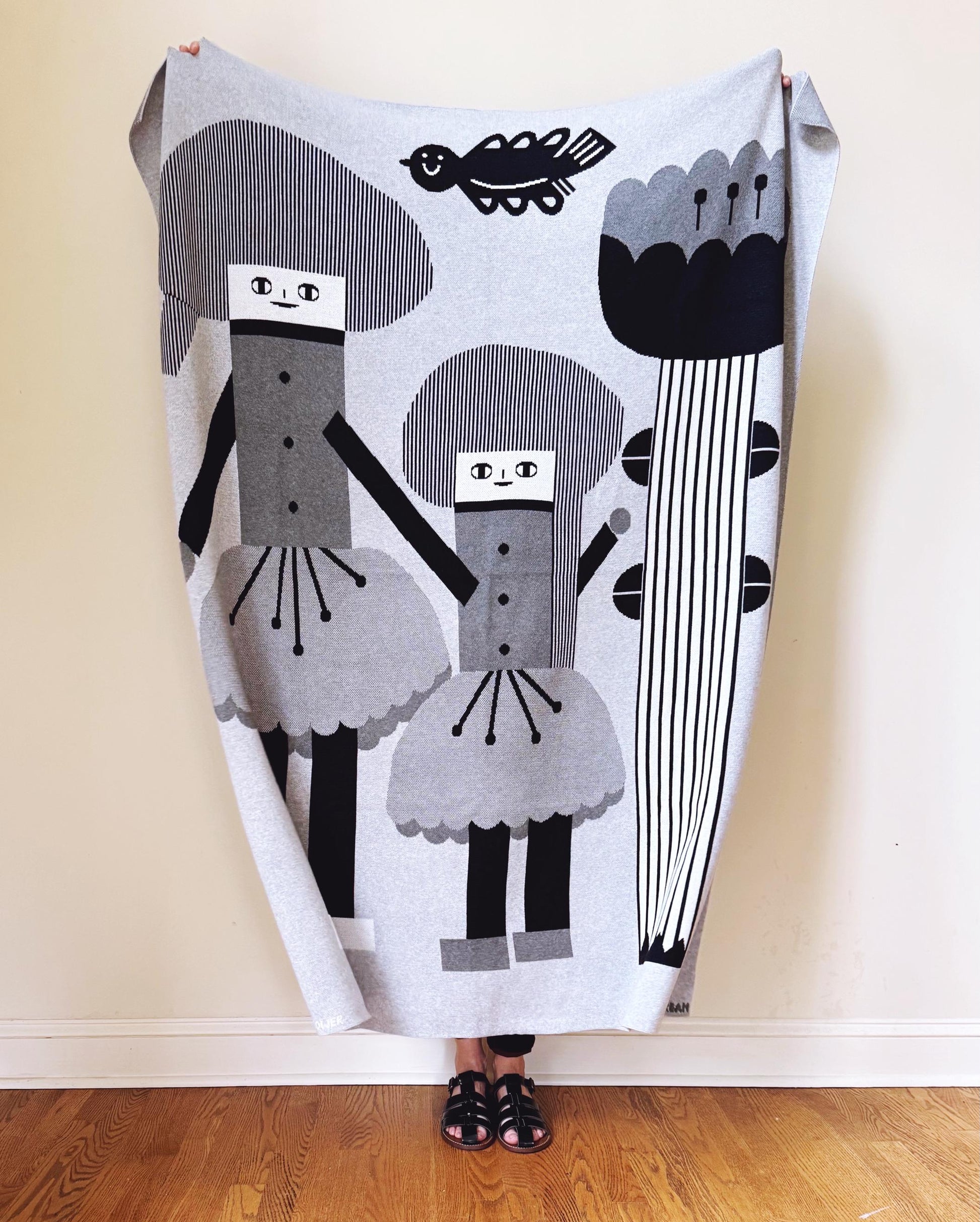 Person holding a decorative knitted blanket with black and gray abstract designs on a wooden floor.
