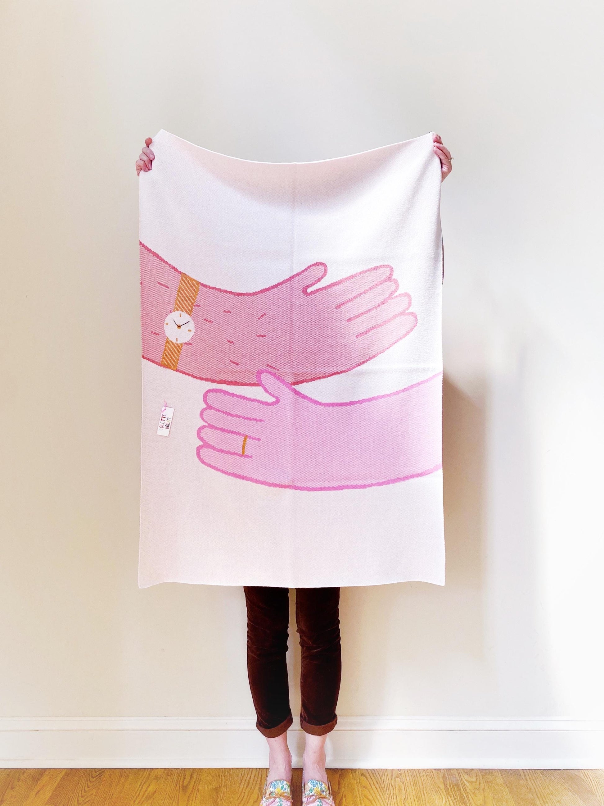 Person holding a knit baby blanket with pink hands in a hug design against a plain background