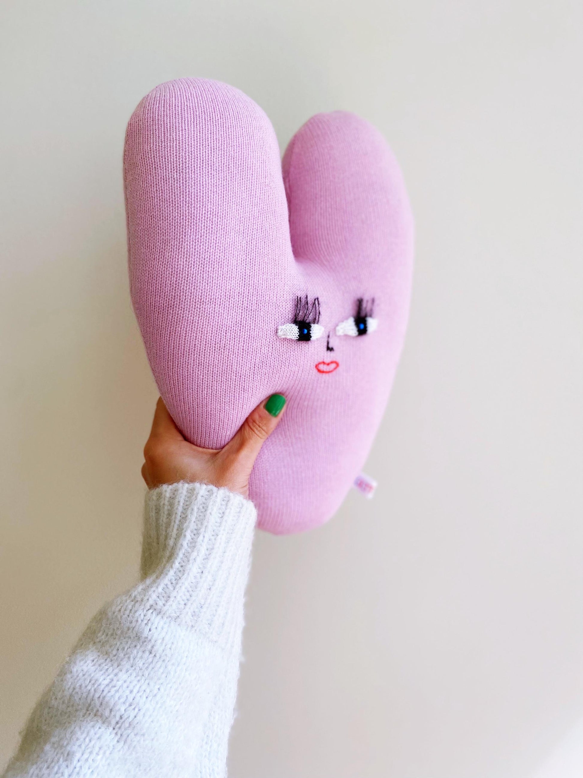 Pink heart-shaped knitted plush toy with face design held by a person against a plain background